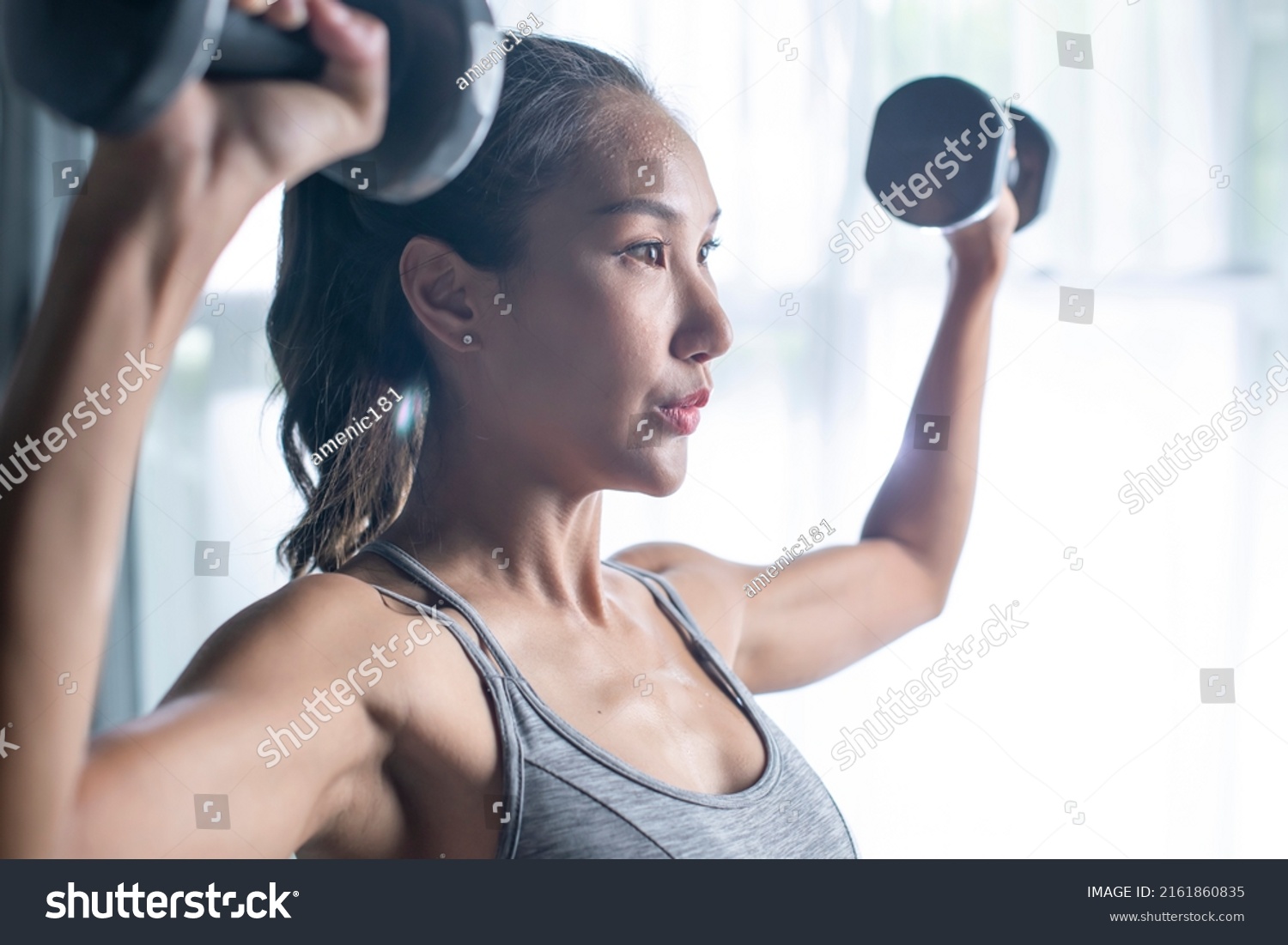Asian young woman doing dumbbell workout at home  working on arms strength  athletic female lifting blue fitness dumbells up over living room interior