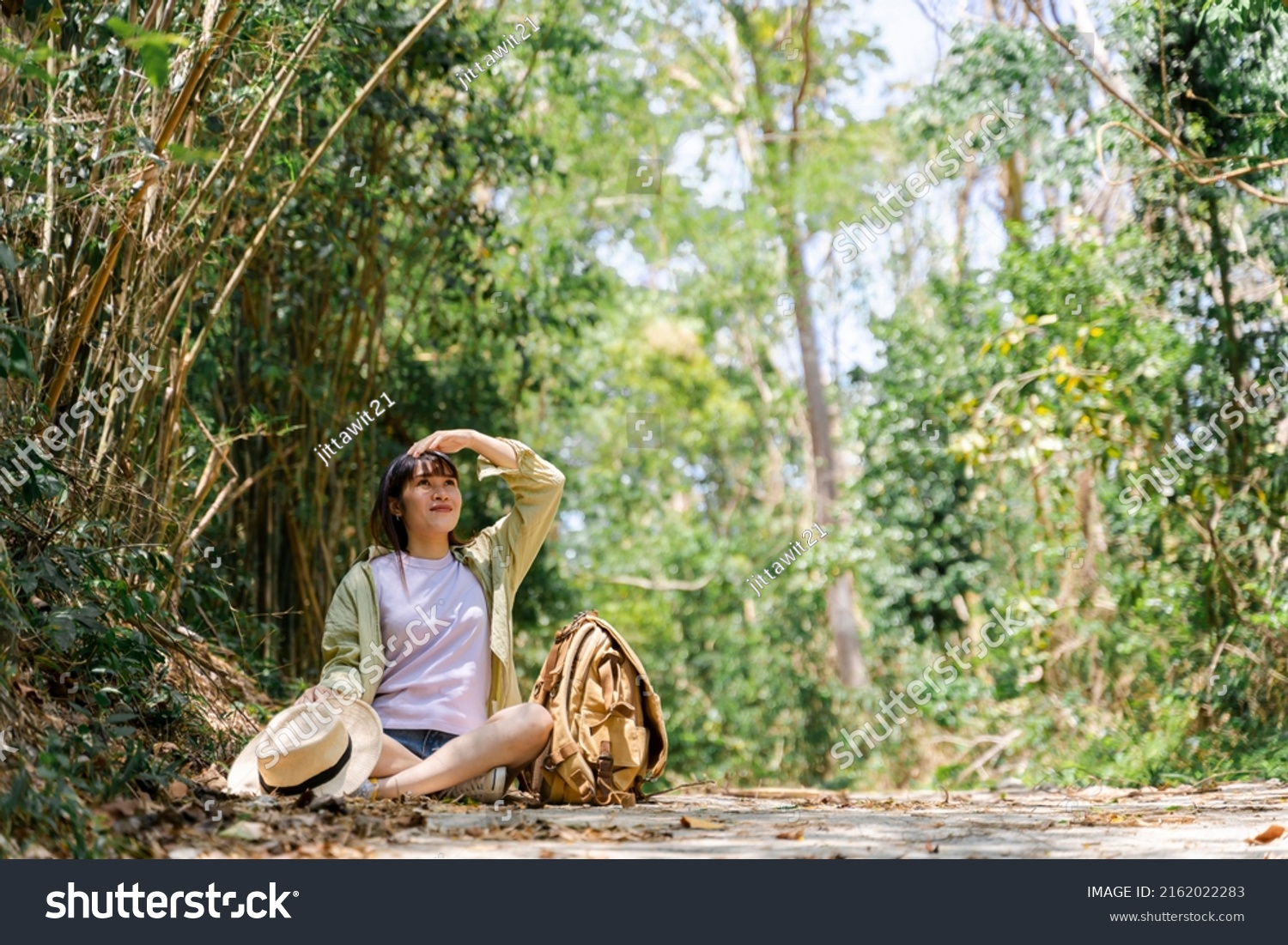 Cute young woman taking a break from forest walk in green tree background with straw fog backpack