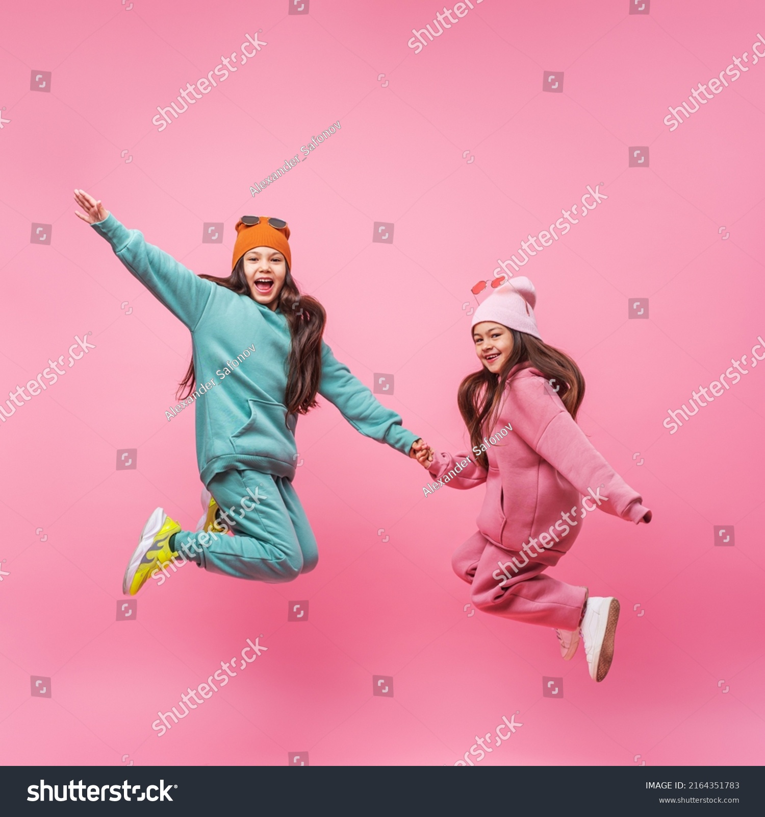 Two smiling Latin child girls in warm fashionable tracksuits jumping on a pink studio copy space. Children's beauty models with dark long hair advertising sports outerwear. Promotion of clothing store