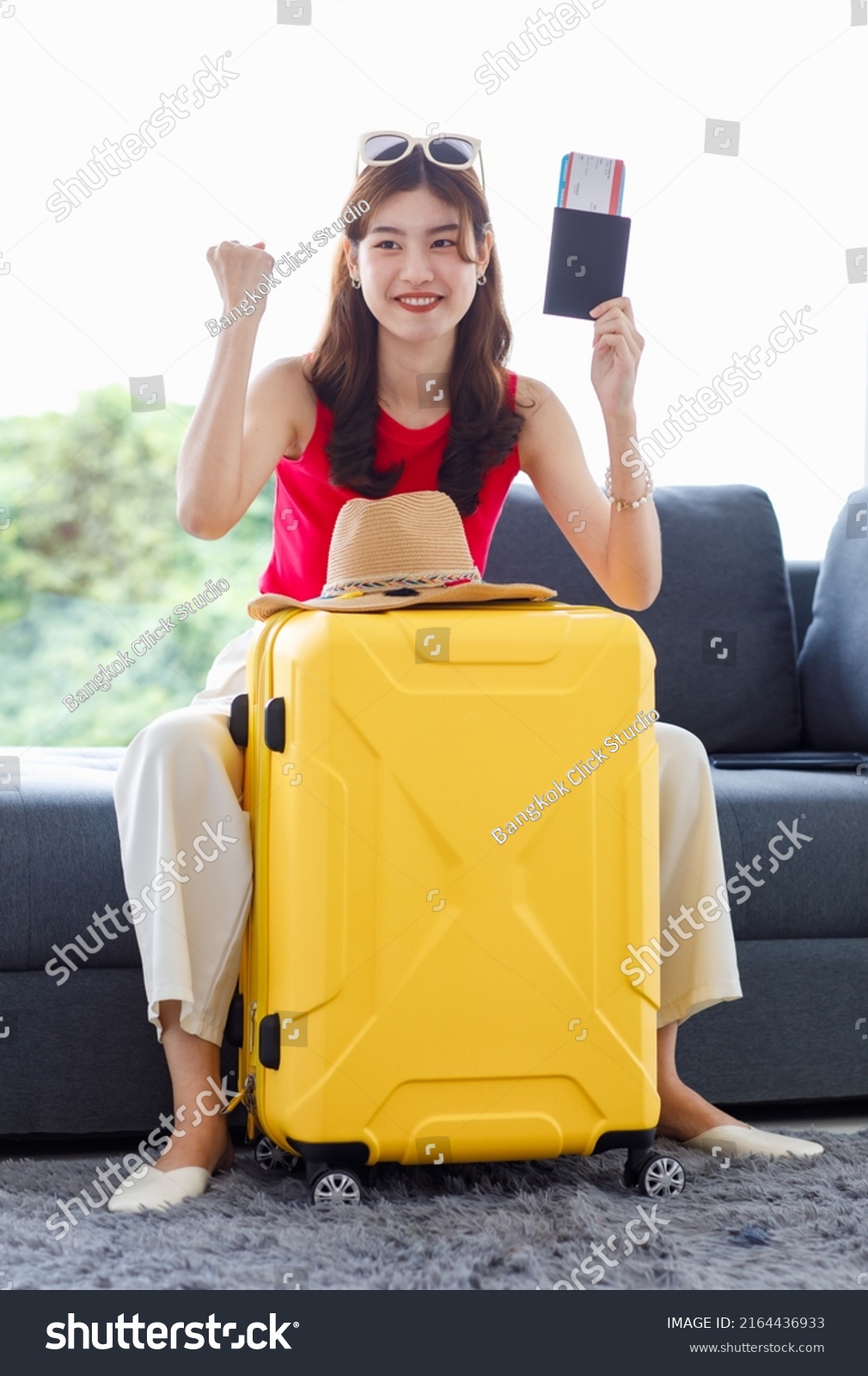 Asian young happy cheerful female traveler in casual summer outfit with sunglasses sitting smiling on cozy sofa in living room holding passport and boarding pass ticket with yellow trolley luggage.