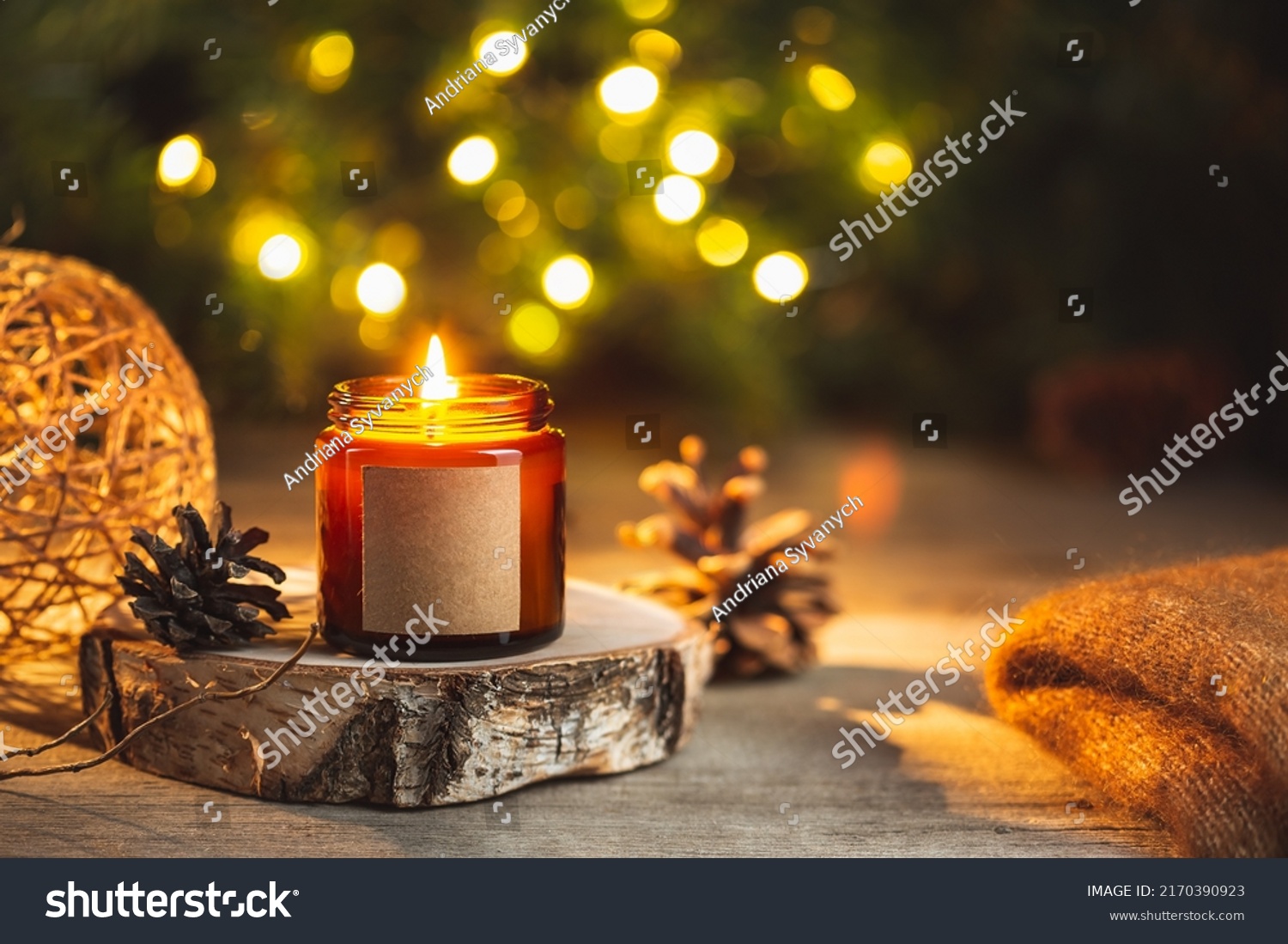 Cozy winter evening card with burning aroma  candle in a dark glass jar with empty label mock up. Soft focus  copy space for text. Blurred bokeh lights on the background. Christmas holiday at home.