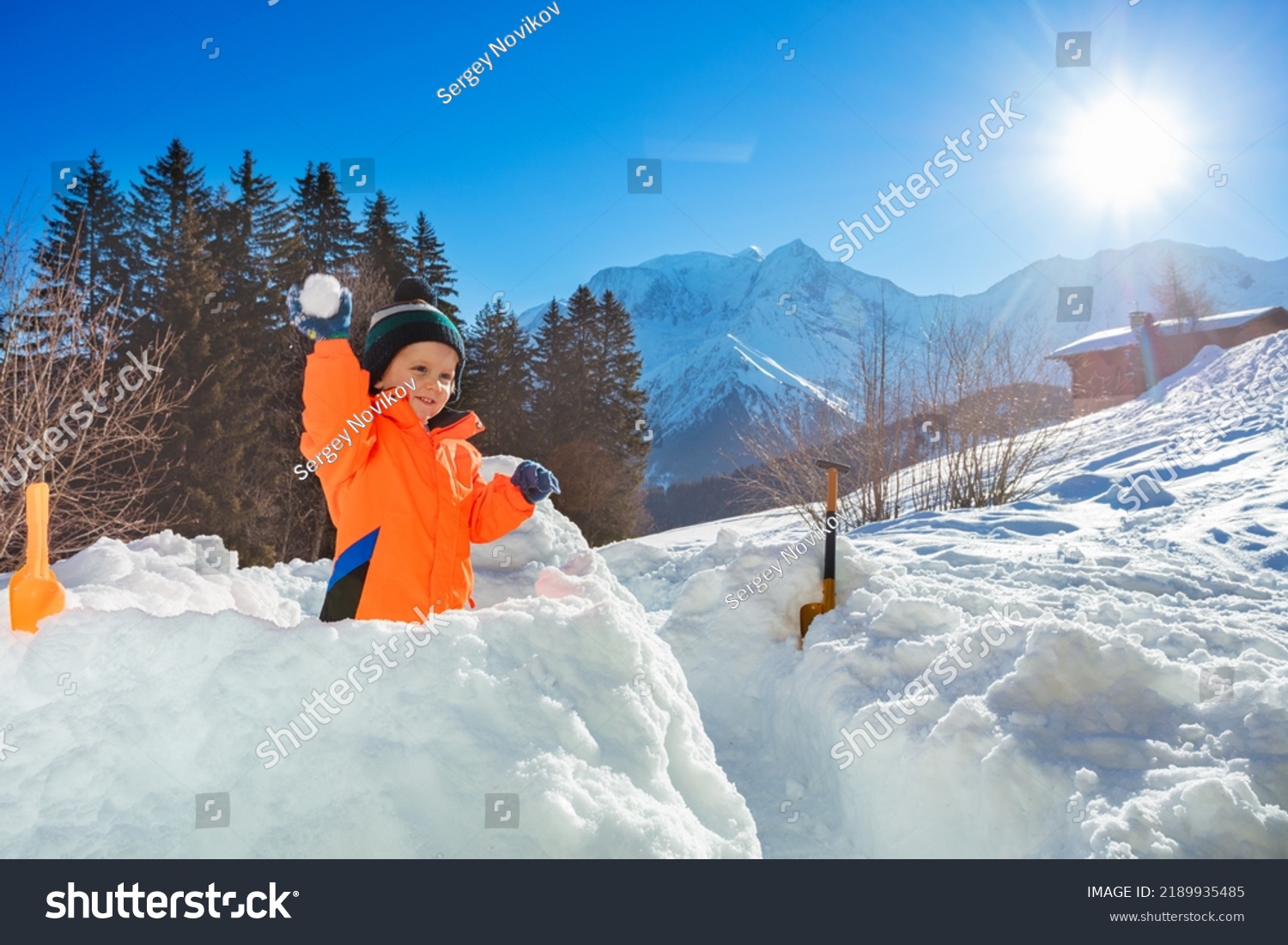 Happy little boy throw snowball standing in the snow fortress with Alps mountains on background during winter vacations