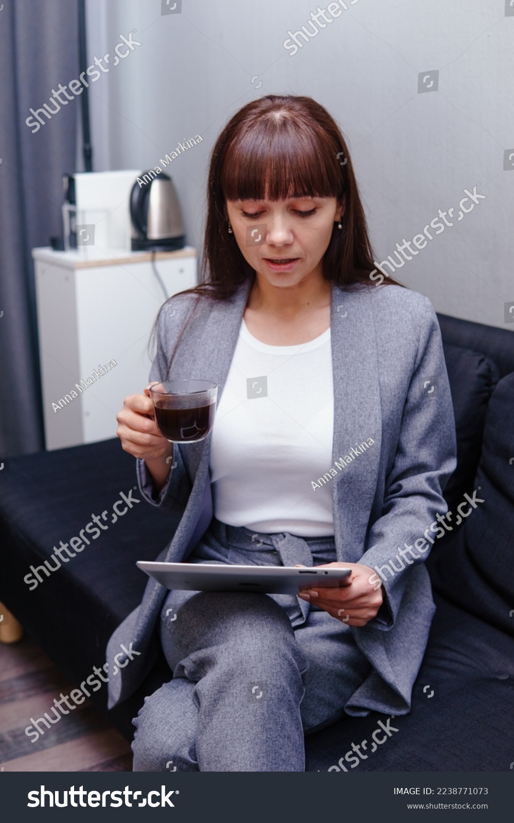 A brunette in a gray suit is sitting on the couch in the office with a cup of black coffee. Business concept. Brunette behind an electronic tablet