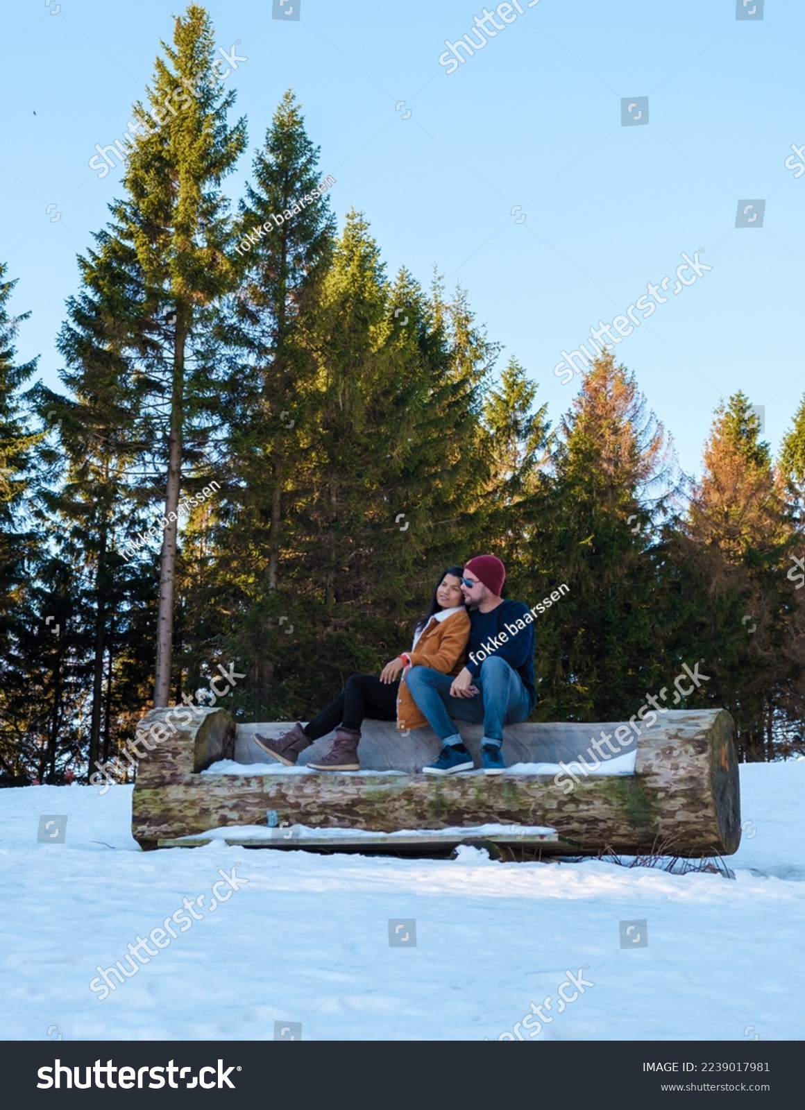 Couple sitting on a wooden bench in the snow Caucasian men and Asian women in the snow with wood in the background at the Harz National park in Germany.