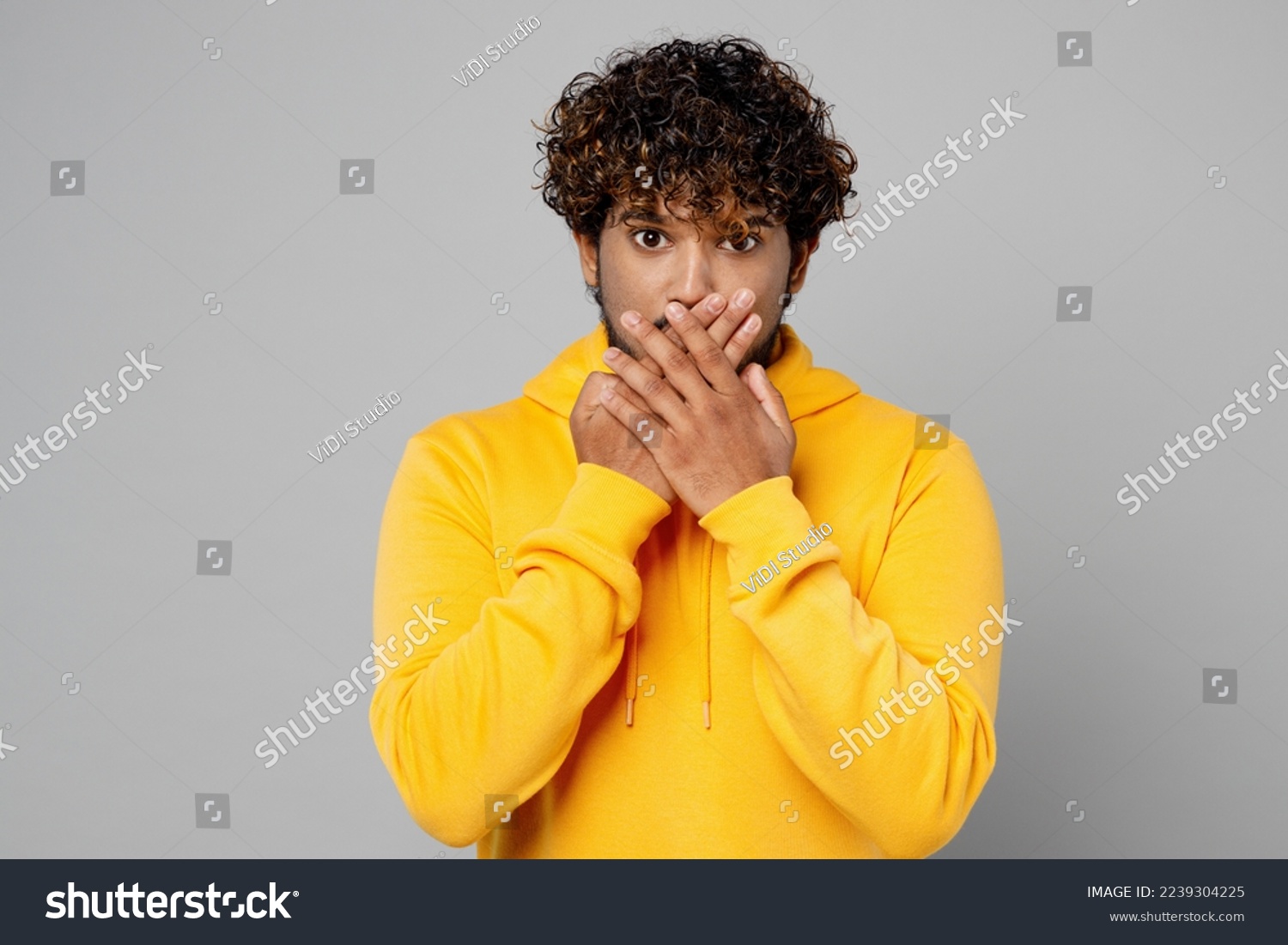 Young amazed shocked scared astonished sad Indian man 20s he wears casual yellow hoody cover mouth with hand look camera isolated on plain grey background studio portrait. People lifestyle portrait