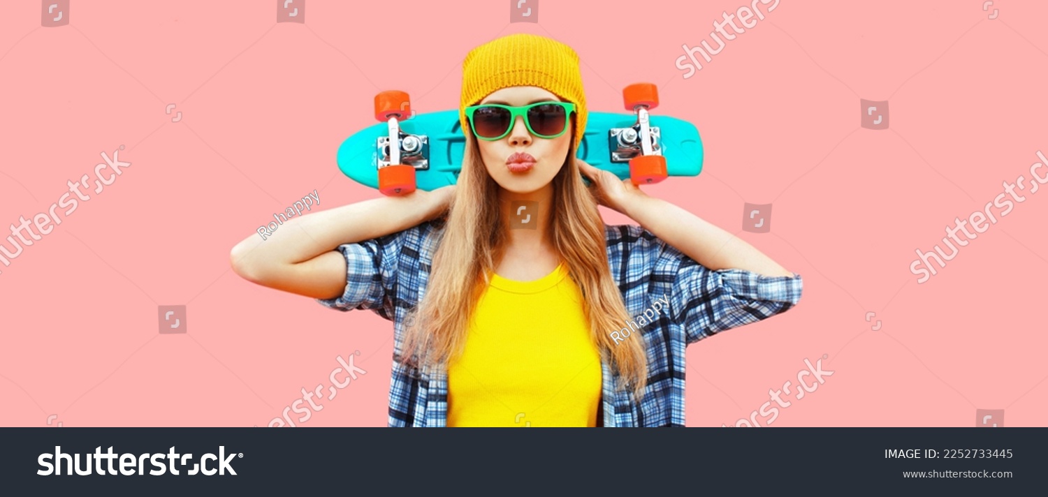 Portrait of stylish blonde young woman model with skateboard wearing colorful yellow hat on pink background