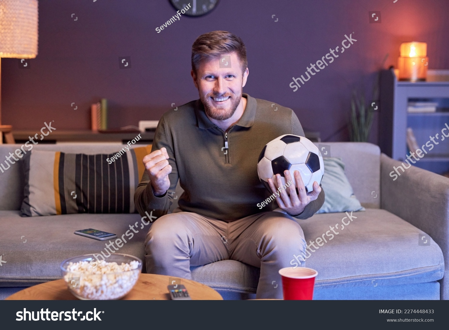 Portrait of emotional man watching sports match on TV at home and holding ball in blue light copy space