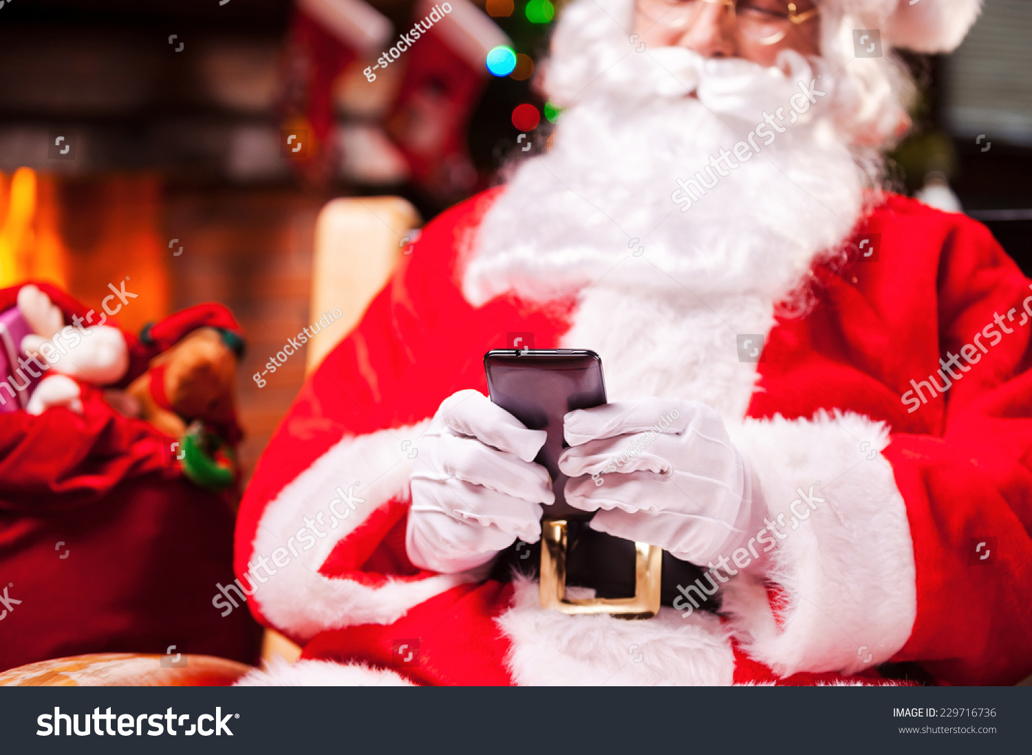 Santa Claus texting. Close-up of Happy Santa Claus typing message on the mobile phone and smiling while sitting at his chair