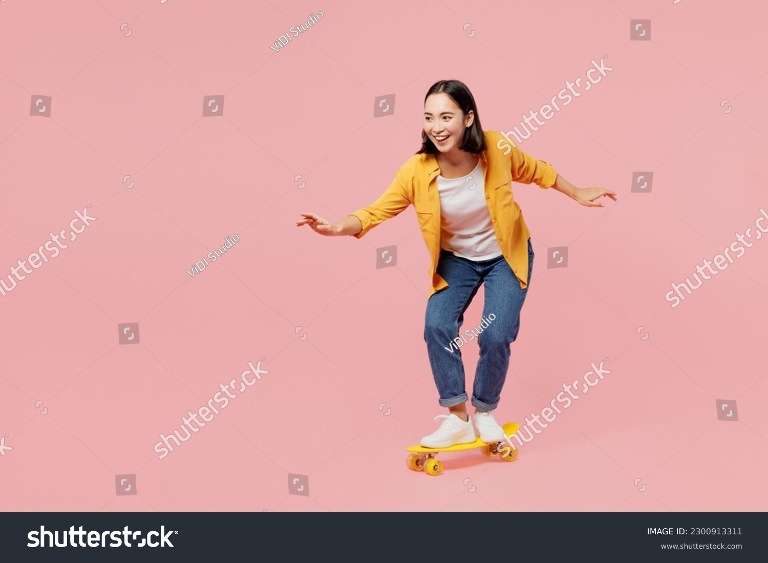Full body young happy fun cool woman of Asian ethnicity wear yellow shirt white t-shirt riding skateboard pennyboard isolated on plain pastel light pink background studio portrait. Lifestyle concept