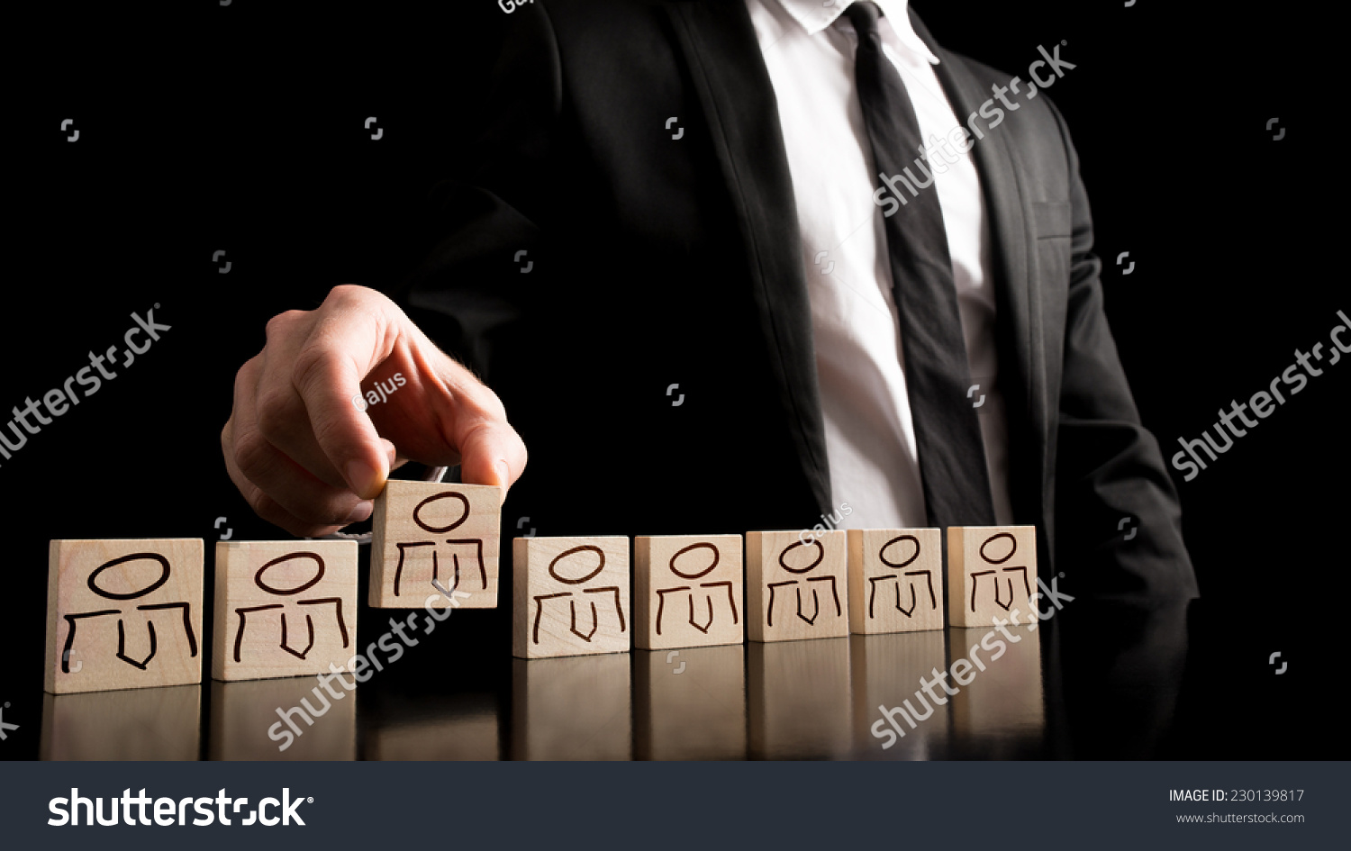 Businessman in black and white business suit arranging wooden pieces with people drawings on the table with pure black background. Simple human resource concept.