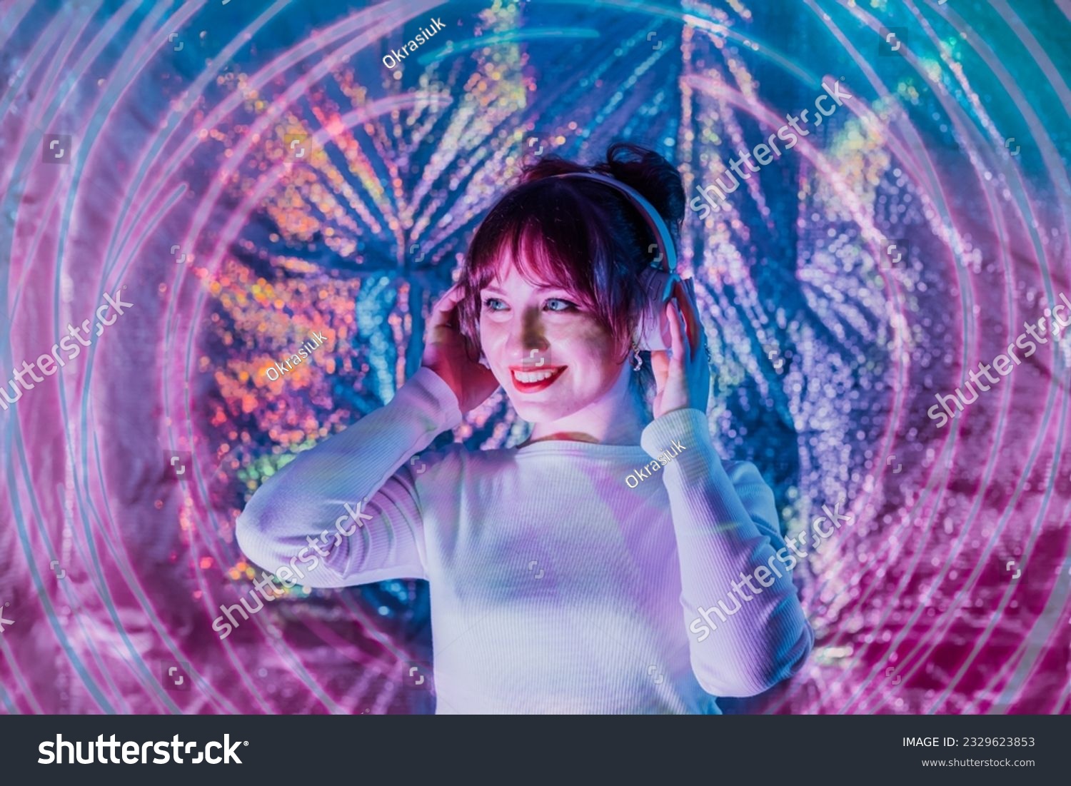 Portrait of relaxed smiling woman in white clothes and headphones dancing in neon light. Music lover. White dress code party. Silent disco. Enjoy moment on shiny background. Selective focus.