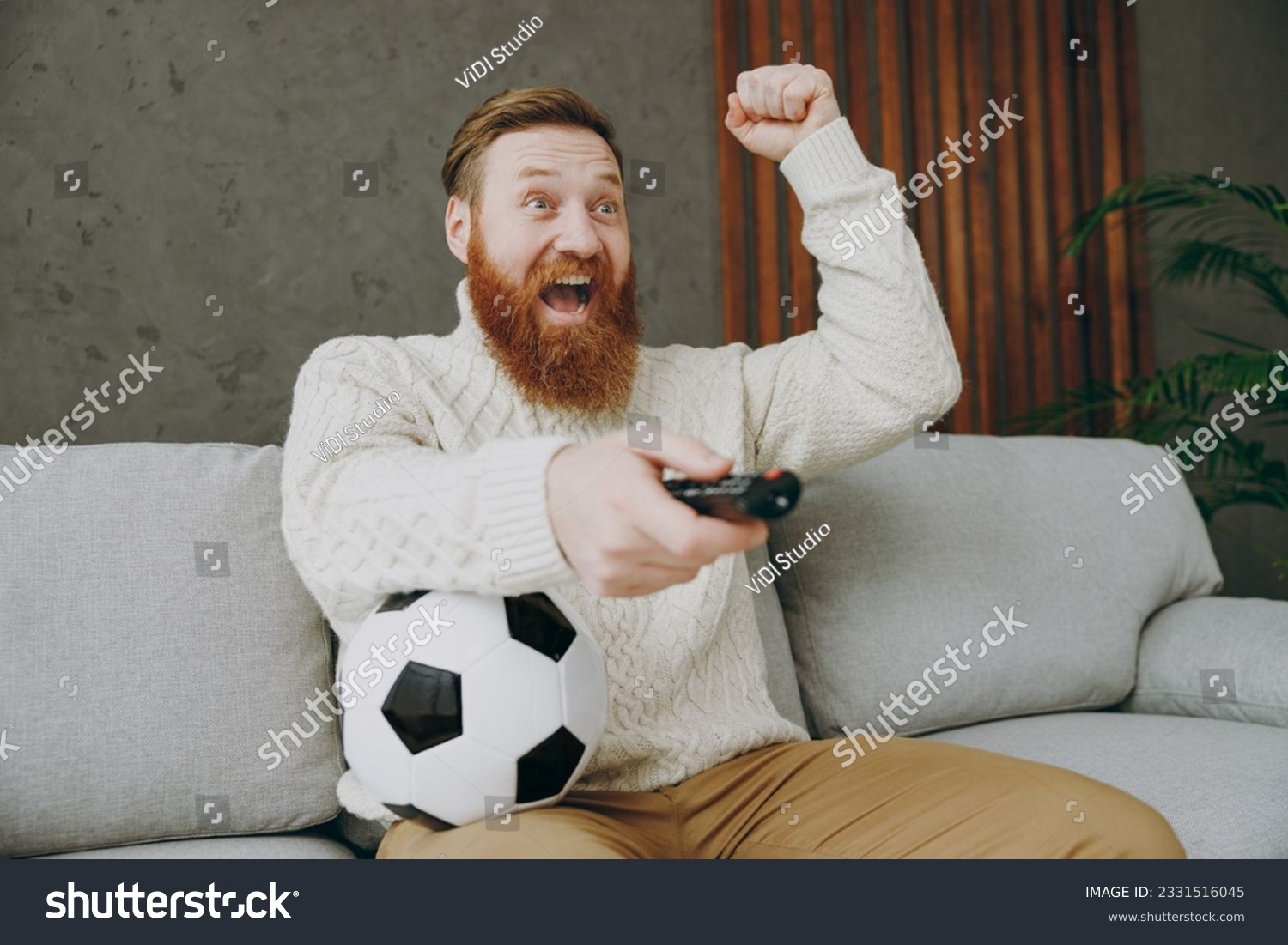 Young man fan wear casual clothes cheer up support football team sit on grey sofa with soccer ball do winner gesture switch channels rest watch tv indoors room gray color wall. Sport leisure concept