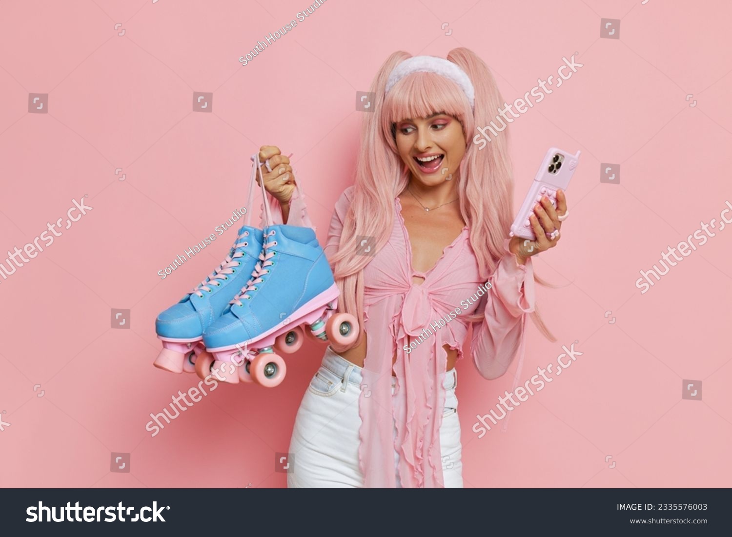 Pretty girl dressed in Barbie style pink clothes with long pink hair holding pink phone and blue roller-skates and smiling on pink background  personal style concept  copy space