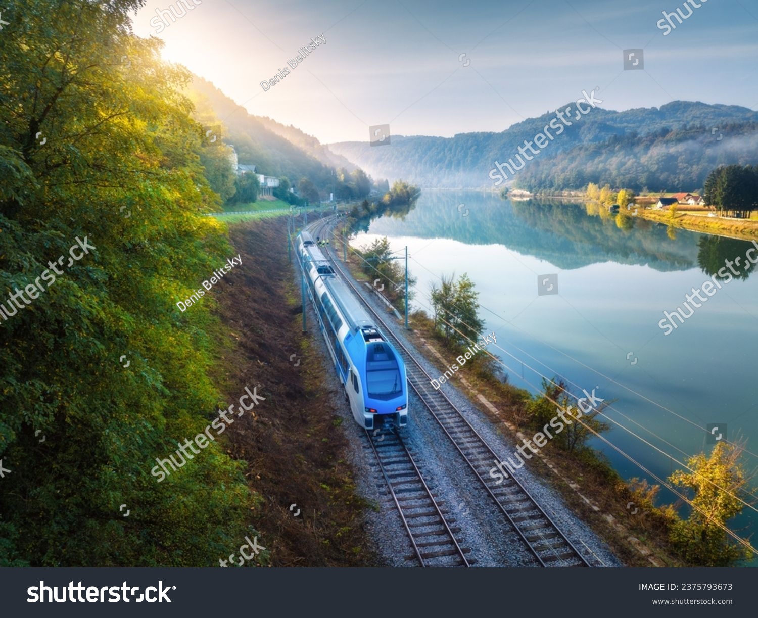 Aerial view of blue modern high speed train moving near river in alpine mountains in fog at sunrise in autumn. Top view of train railroad lake reflection trees in fall. Railway station in Slovenia