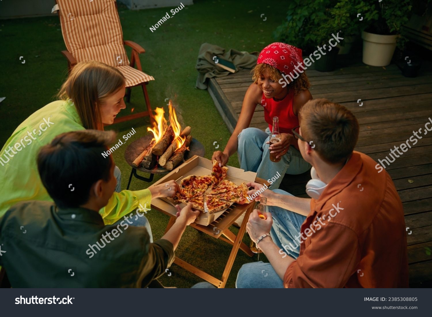 Happy friends enjoying pizza and beer drink nearby burning campfire