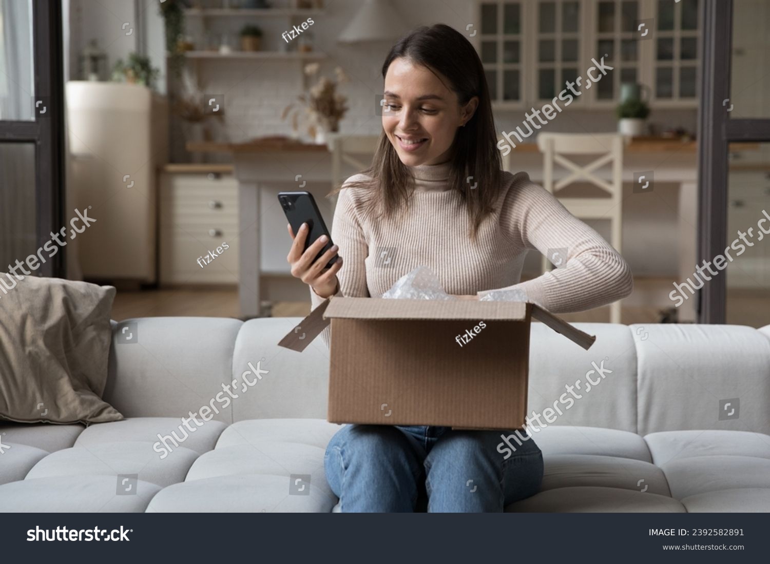 Pretty woman sit on sofa opens received courier parcel box reviewing order in retail services using app on smart phone check items feels happy with delivered goods. E-commerce client tech concept