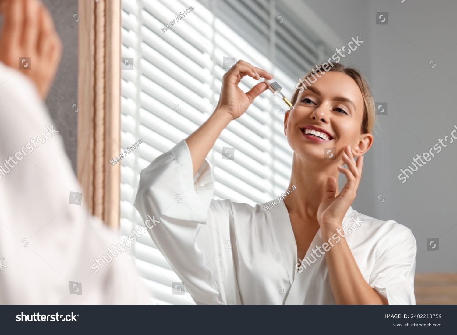 Beautiful woman applying cosmetic serum onto her face near mirror in bathroom