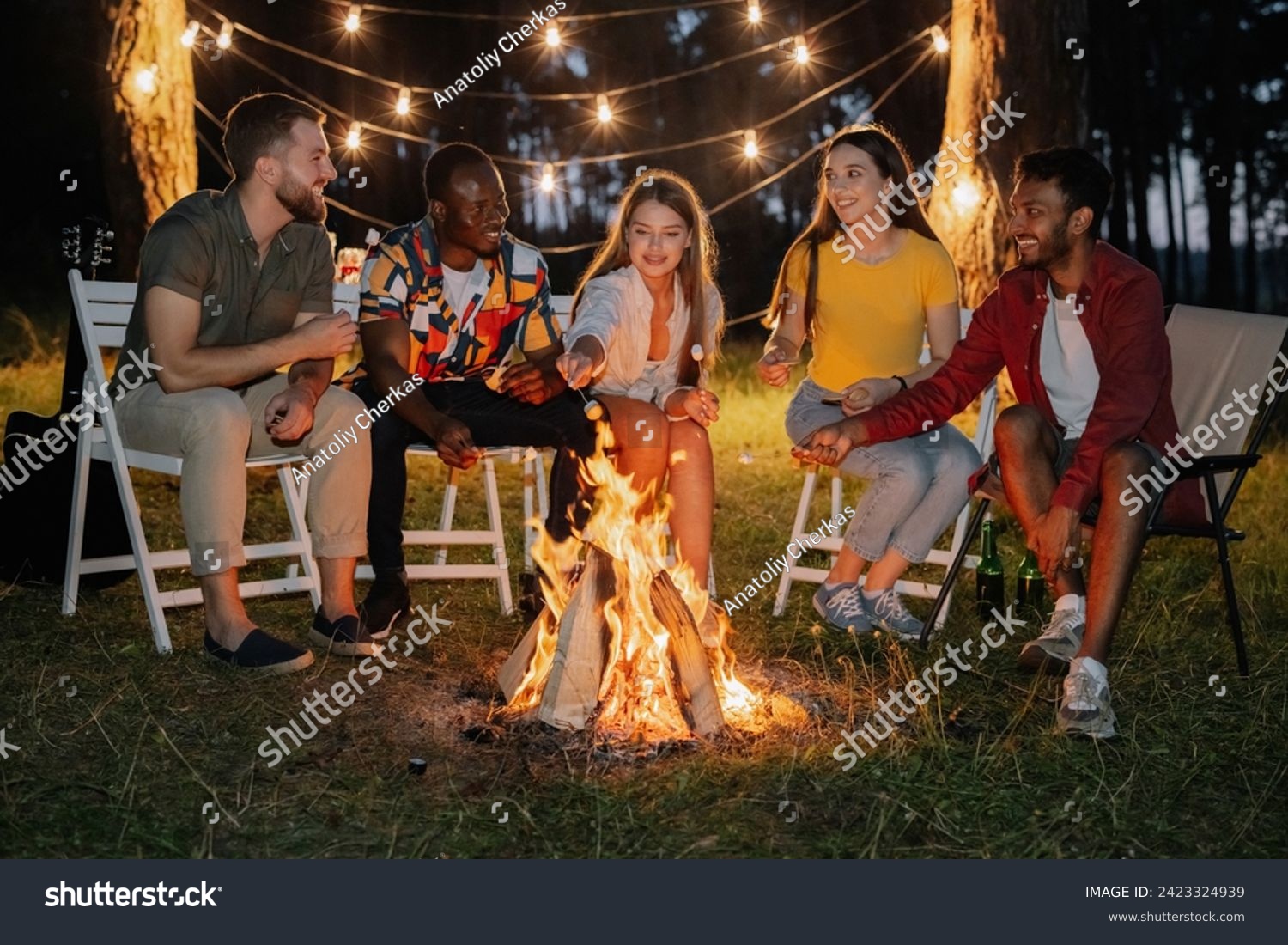 Group of multiracial restful friends roasting marshmallows while sitting by bonfire