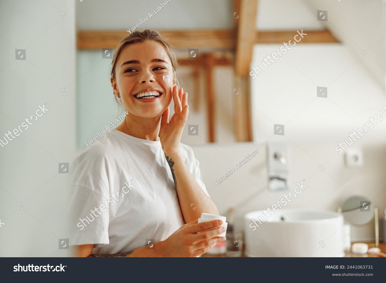 Beautiful young woman taking care of skin by applying moisturizer cream in bathroom