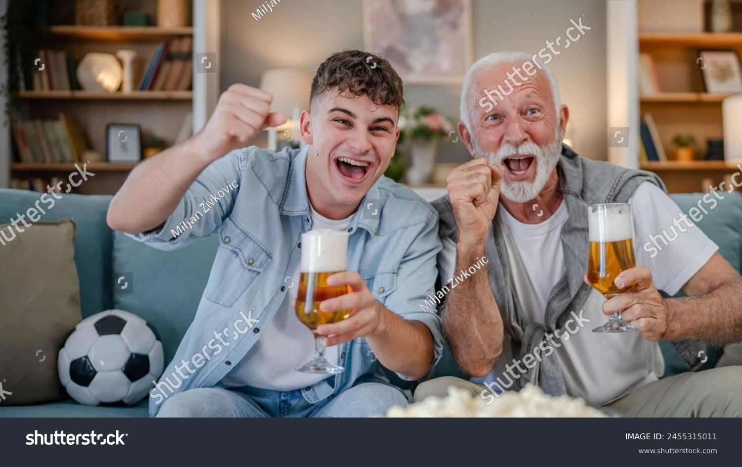 Caucasian teenager and senior man grandfather and grandson sit on sofa bed at home watch football soccer game hold popcorn bowl and glass of beer have fun family bonding males men