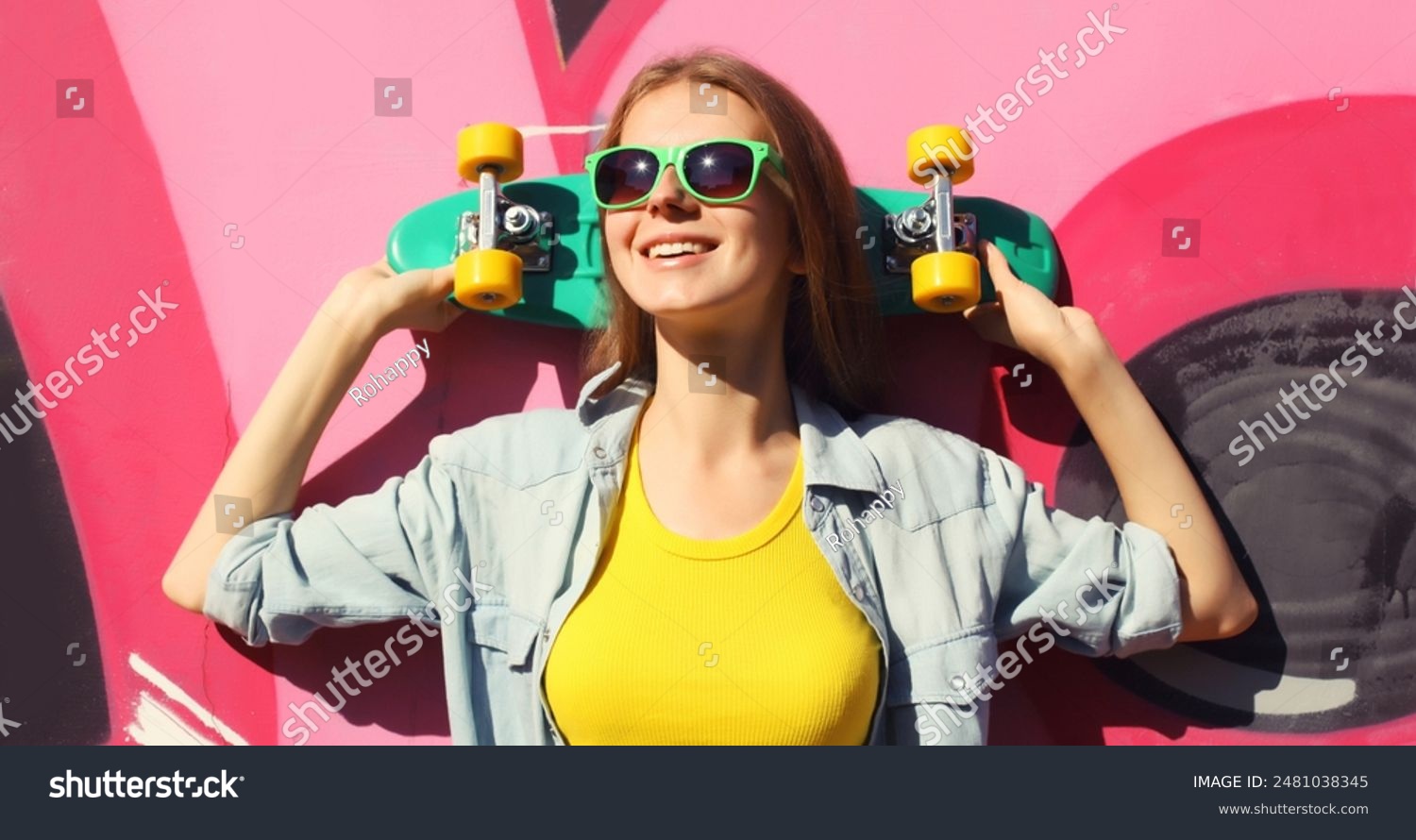 Stylish happy young girl with skateboard in glasses  modern teenager smiling girl looking away posing against the background of pink graffiti wall