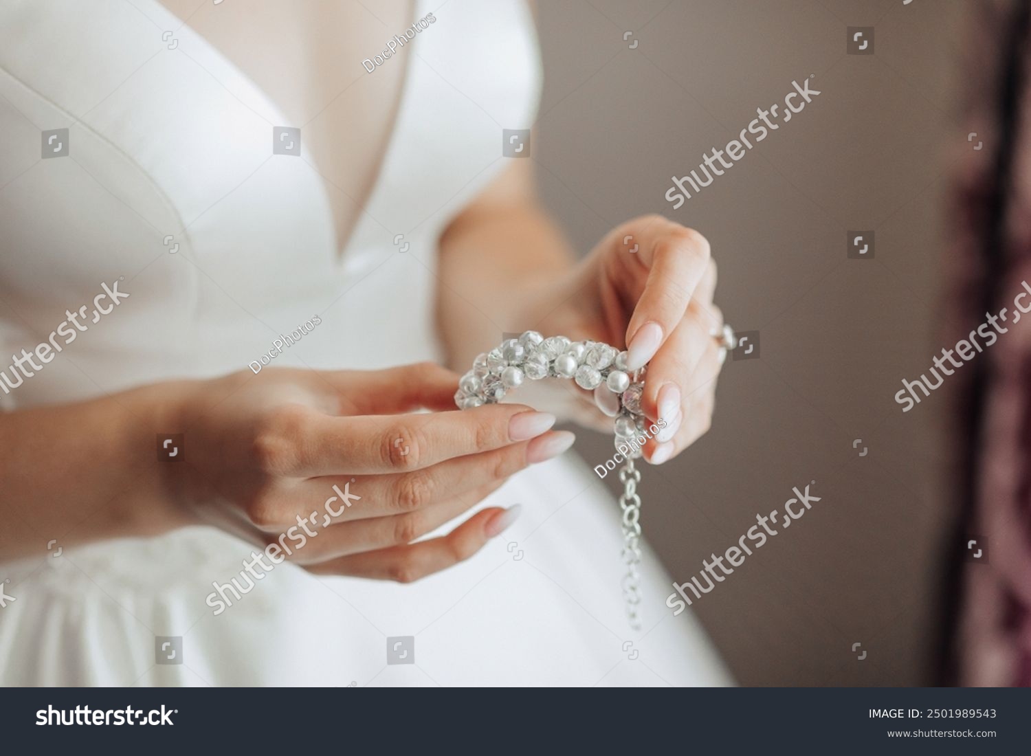 A woman is holding a necklace with pearls and diamonds. The necklace is white and has a gold chain. The woman is wearing a white dress and is standing in front of a curtain