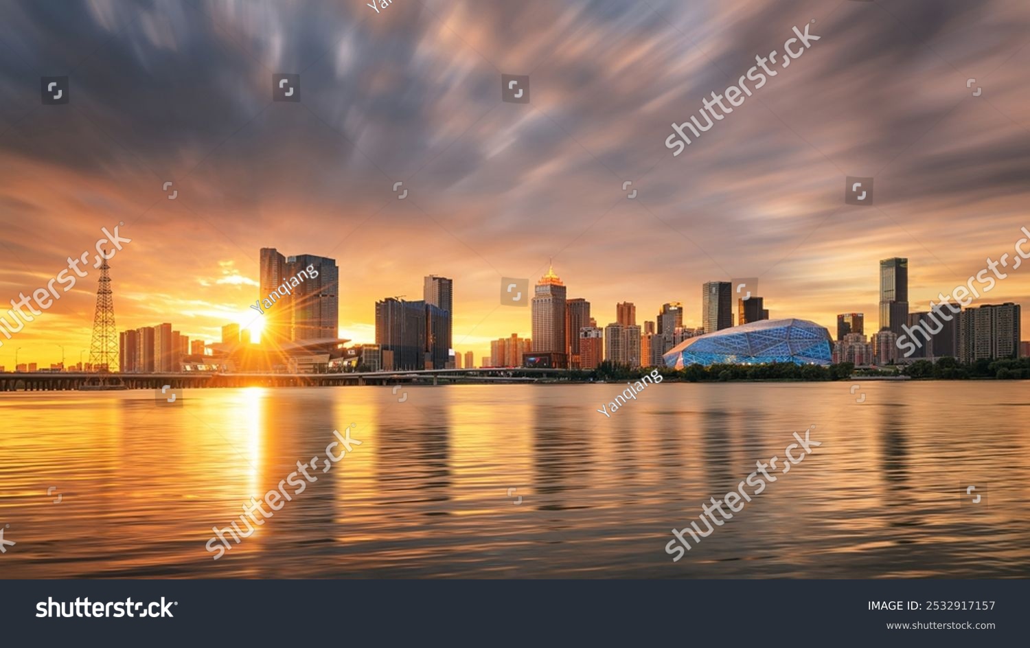 Sun setting behind a modern skyline river reflecting golden hues with dramatic clouds and a geometric blue building adding a futuristic touch.