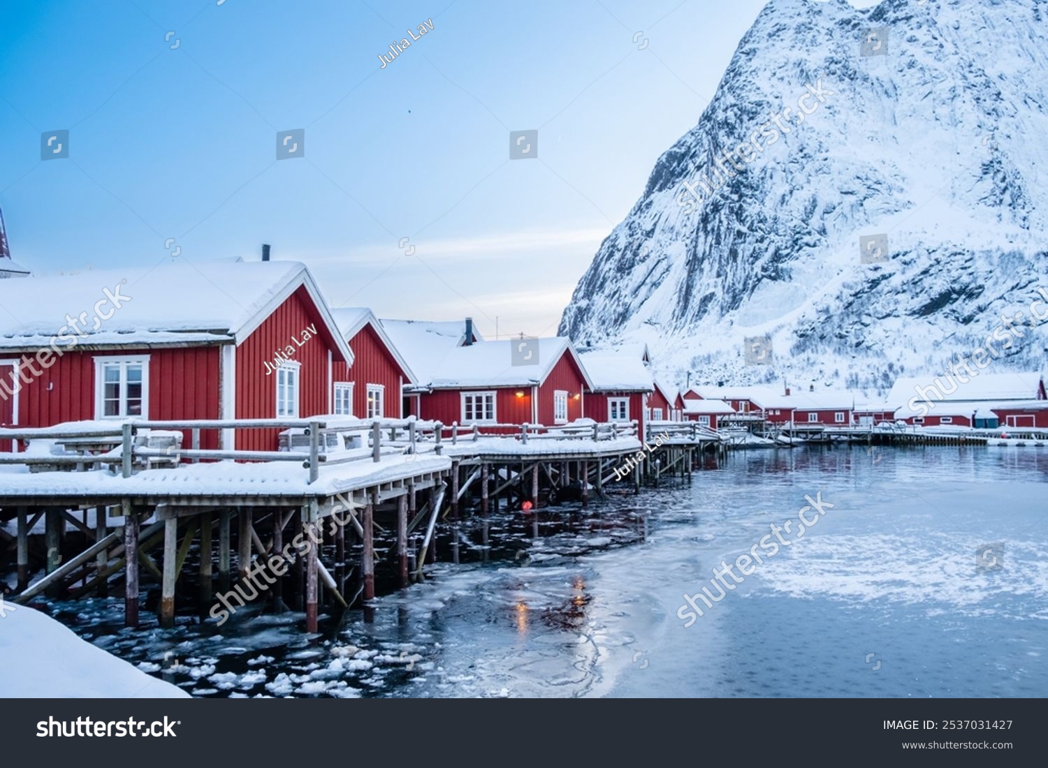 Reine village with traditional rorbu houses on Lofoten islands in winter. Scandinavian landscape with red wooden fishermen cabins on frozen sea water and snowy Reinebringen mountain northern Norway.