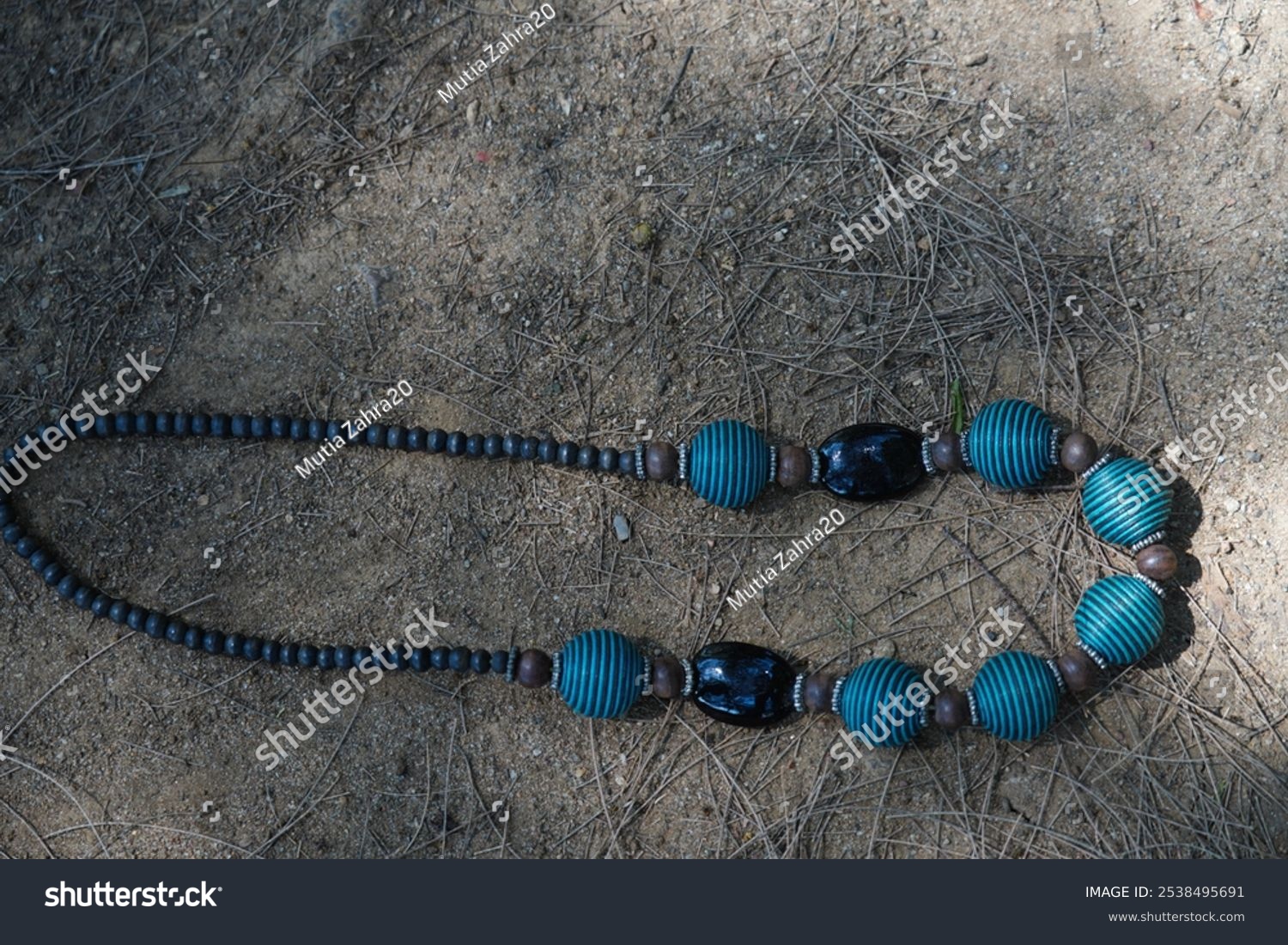 black and blue beaded necklace with round and textured shape  lies on sandy ground with dry twigs.