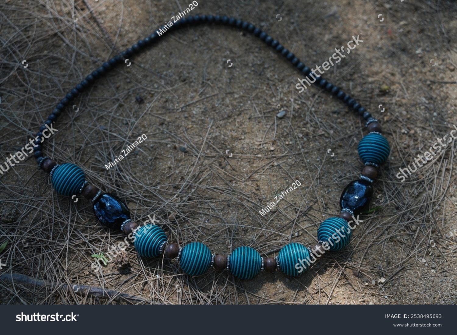 black and blue beaded necklace with round and textured shape  lies on sandy ground with dry twigs.