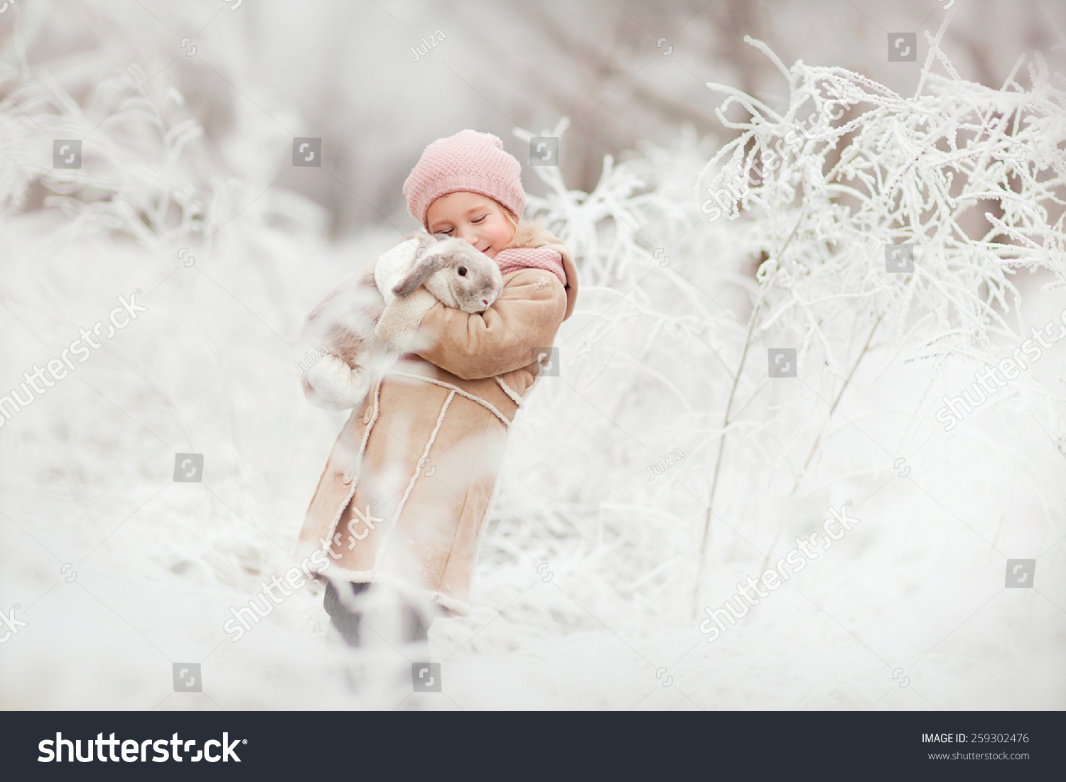 little cute smiling girl in pink hat and beige coat standing in the winter magic forest and holding a rabbit in her hands