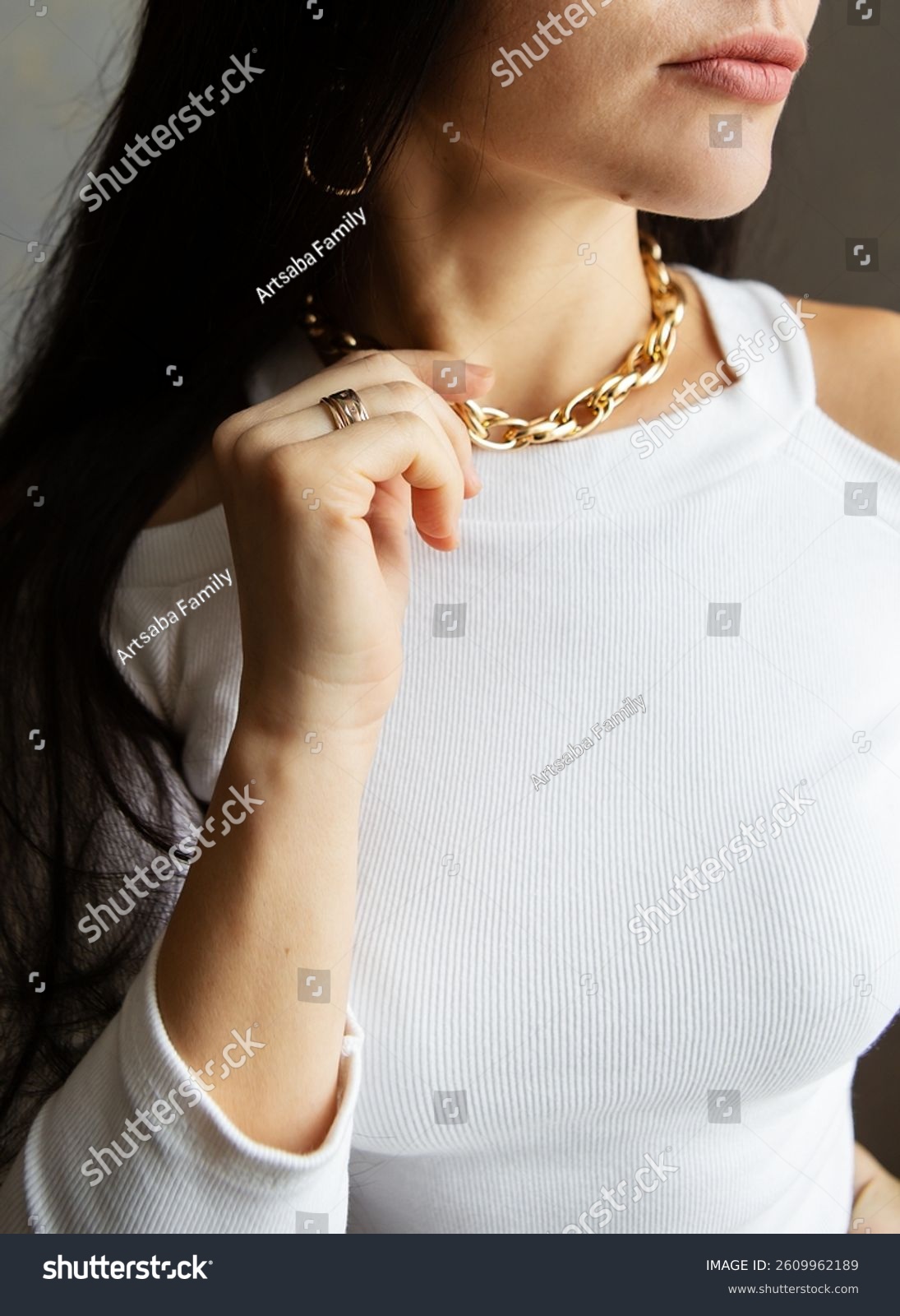 A stylish woman with long dark hair models a gold chain necklace and ring  showcasing elegant jewelry against a white ribbed top.