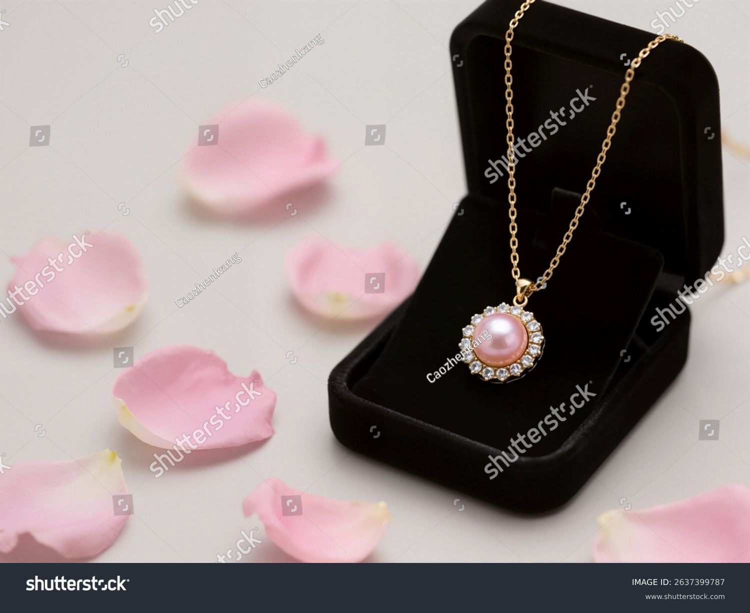 A beautiful pink pearl pendant necklace  set in gold with small sparkling stones  rests in a black velvet gift box. Pink rose petals and a large rose adorn the background.