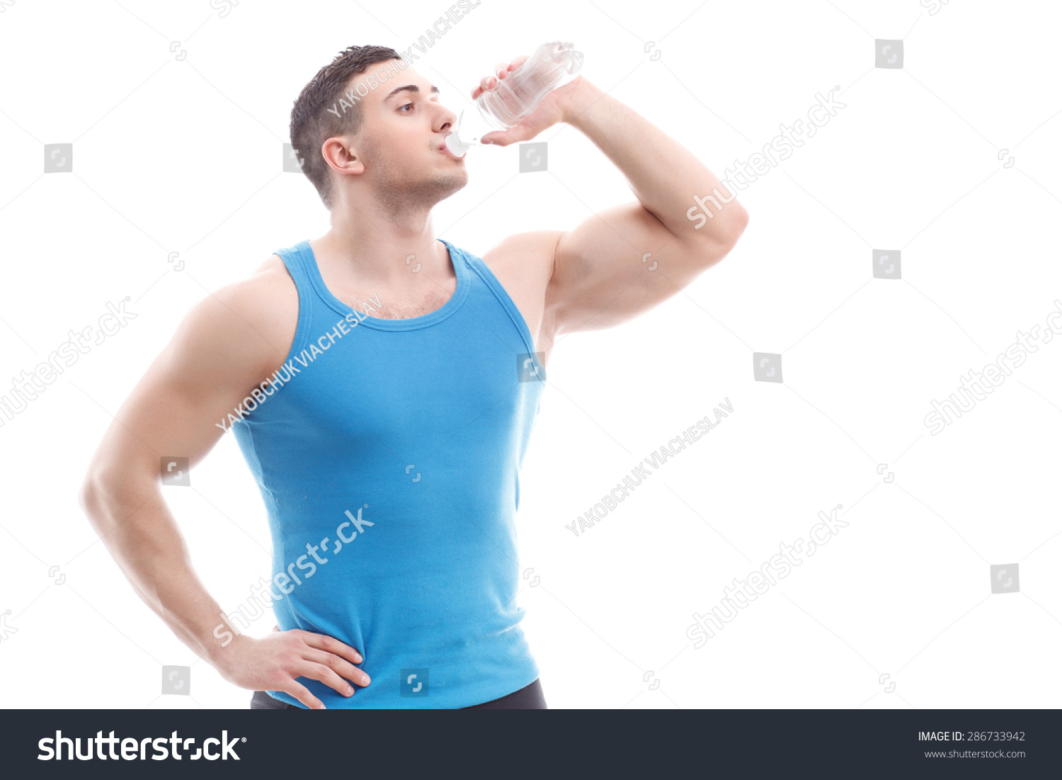 Portrait of a handsome muscled sportsman wearing blue shirt standing smiling holding one hand on his waist and drinking still water from the bottle isolated on a white background