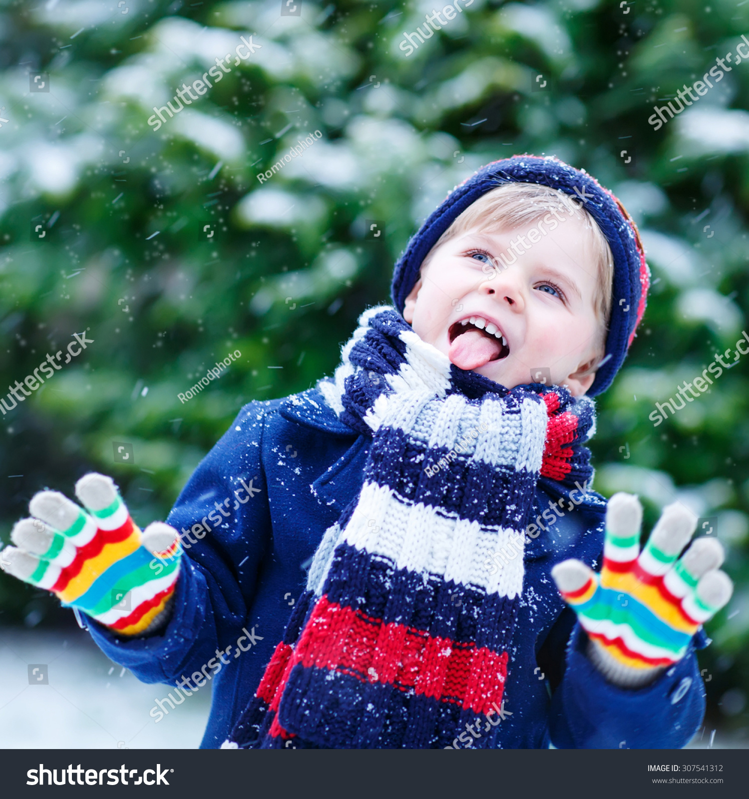 Cute little funny child in colorful winter clothes having fun with snow outdoors during snowfall. Active outdoors leisure with children in winter. Kid with hat hand gloves with stripes.