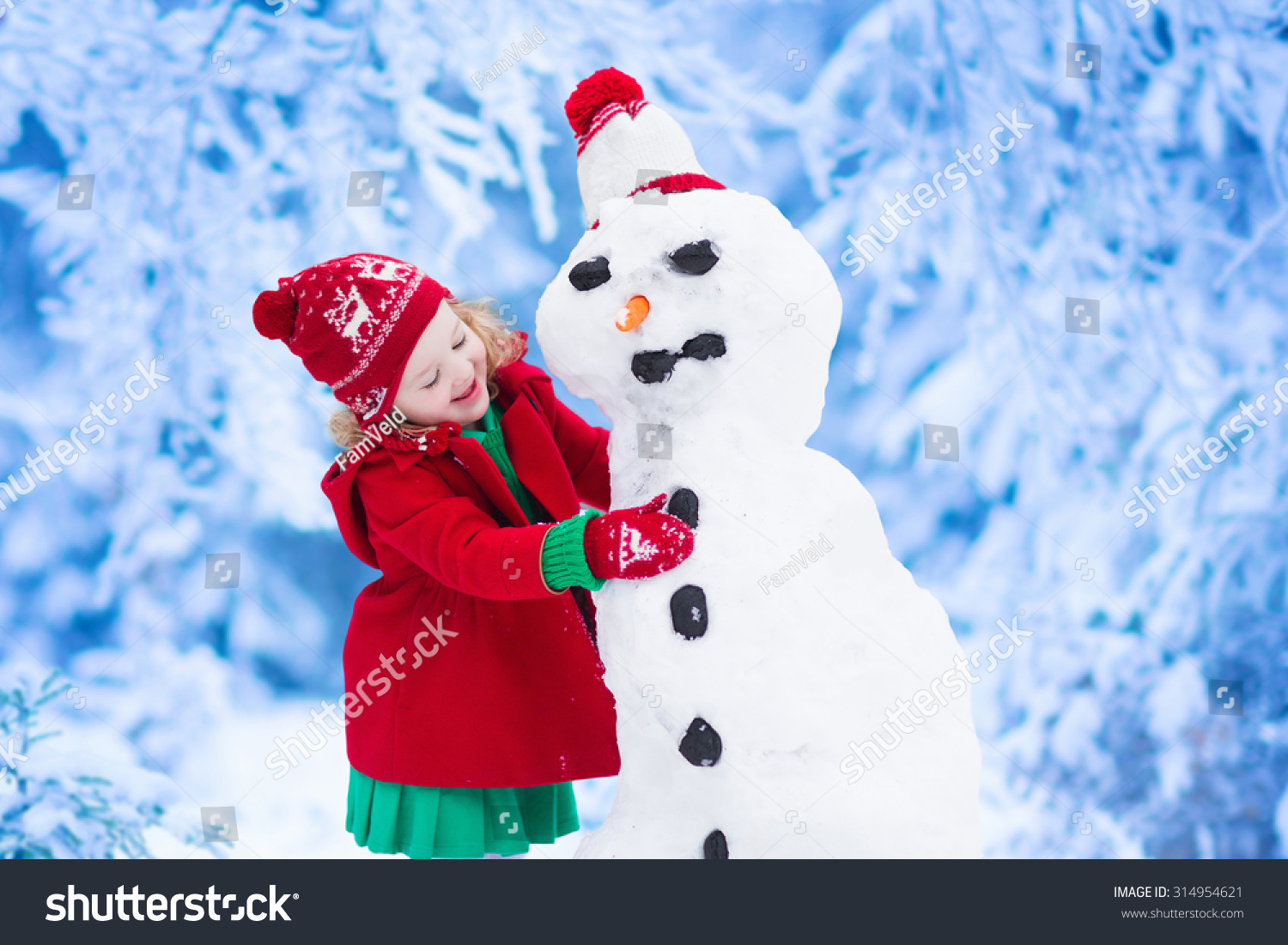 Funny little toddler girl in a red knitted Nordic hat and warm coat playing with a snow man. Kids play outdoors in winter. Children having fun at Christmas time. Child building snowman at Xmas.