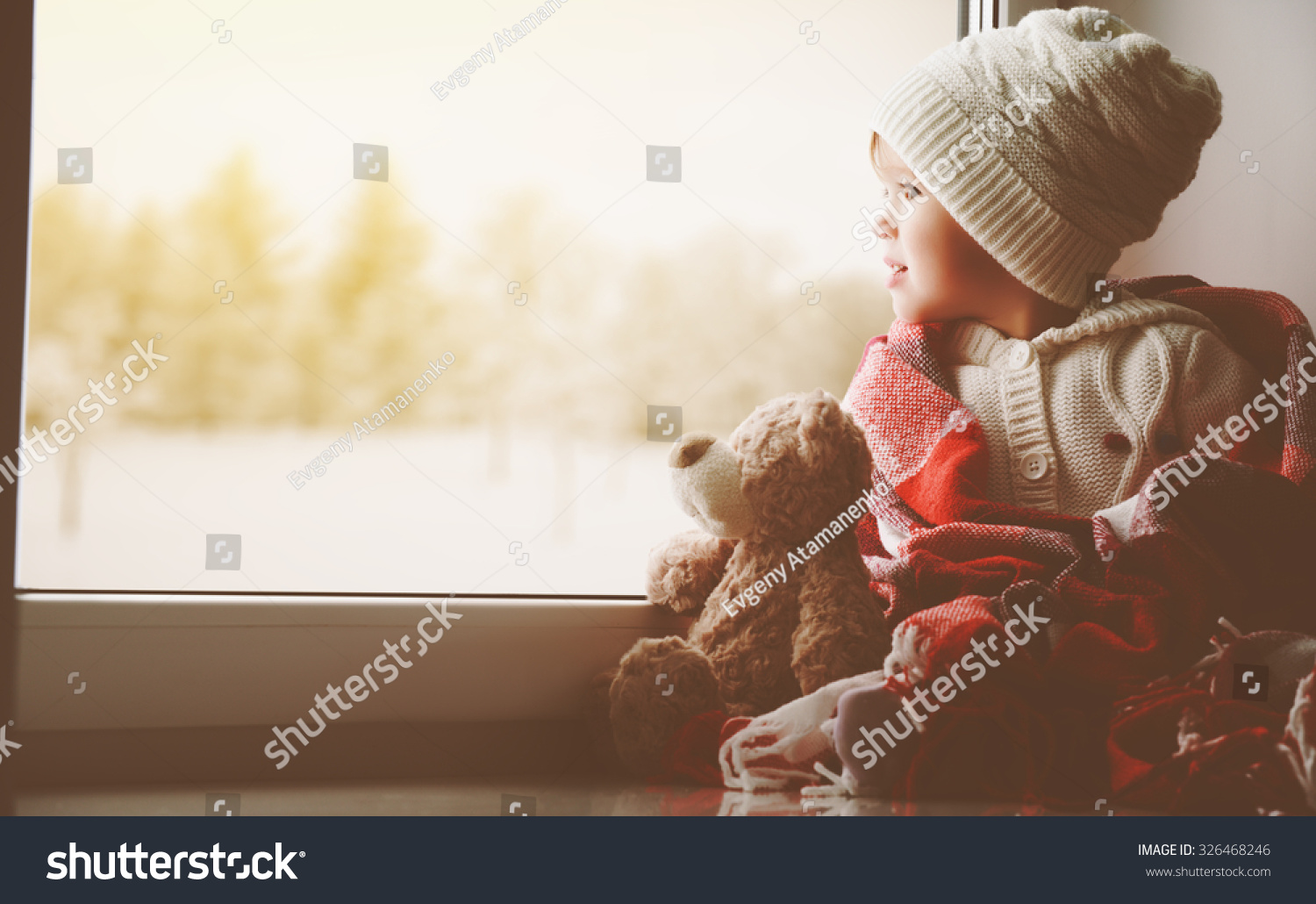child little girl sitting by the window with a teddy bear and looking at the winter forest