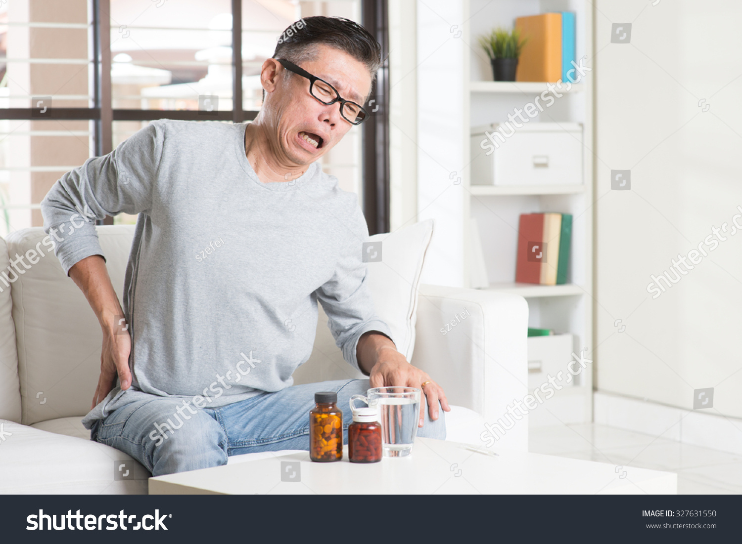 Portrait of casual 50s mature Asian man back pain pressing on hip with painful expression sitting on sofa at home medicines and water on table.