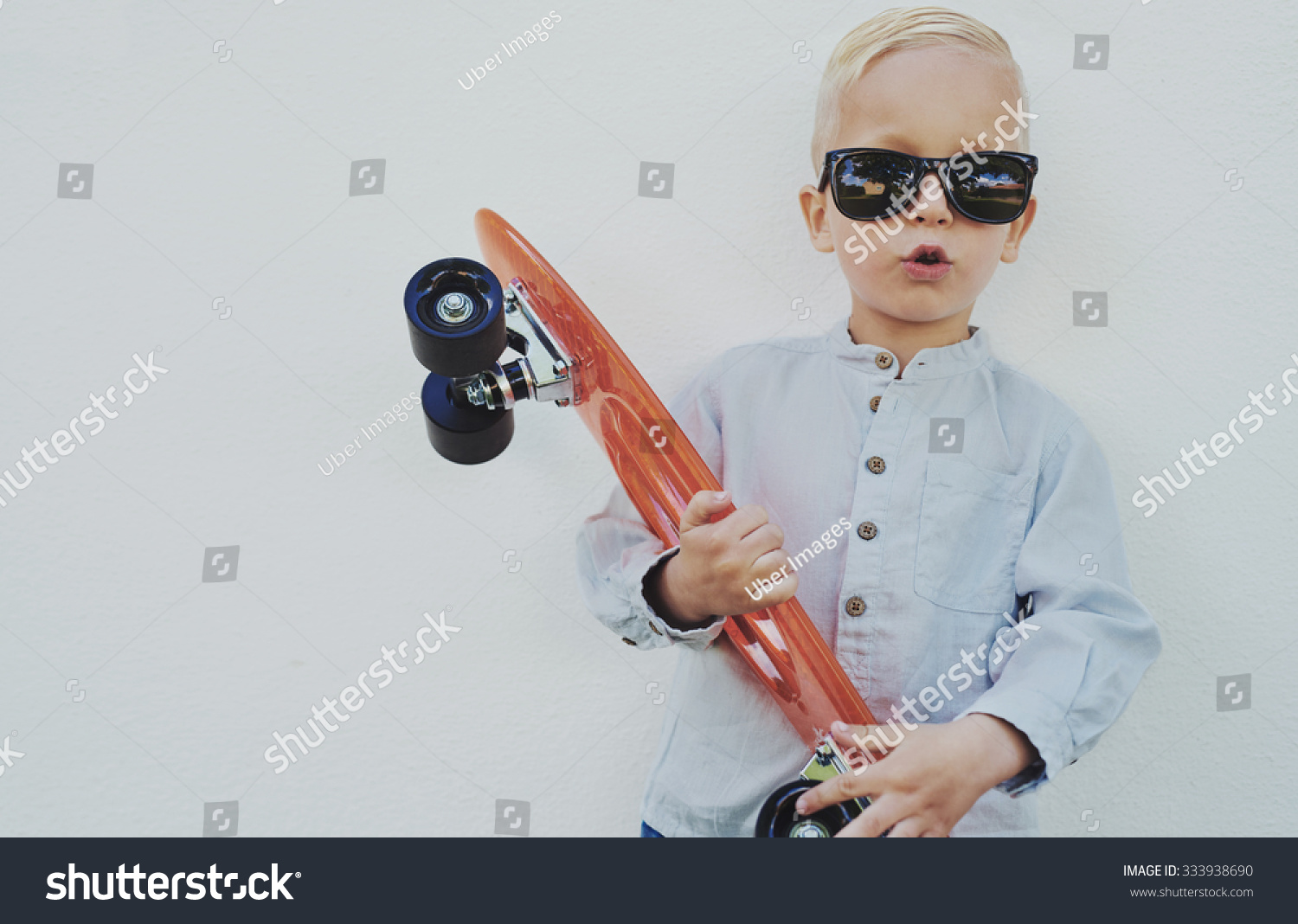 Adorable hipster little boy with a skateboard wearing borrowed sunglasses from his Mum or Dad standing against a wooden wall giving the camera a funny cute expression