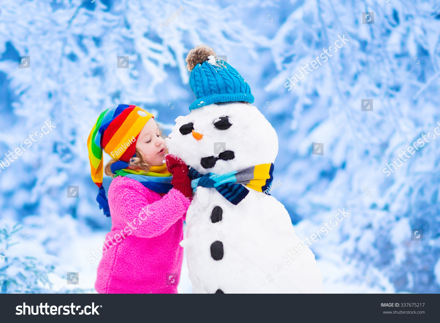 Funny little toddler girl in a colorful hat and warm coat playing with a snow man. Kids play outdoors in winter. Children having fun at Christmas time. Child building snowman at Xmas.