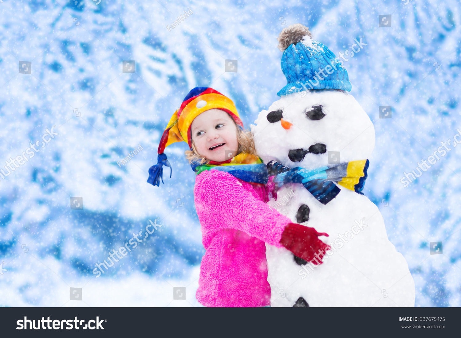 Funny little toddler girl in a colorful hat and warm coat playing with a snow man. Kids play outdoors in winter. Children having fun at Christmas time. Child building snowman at Xmas.