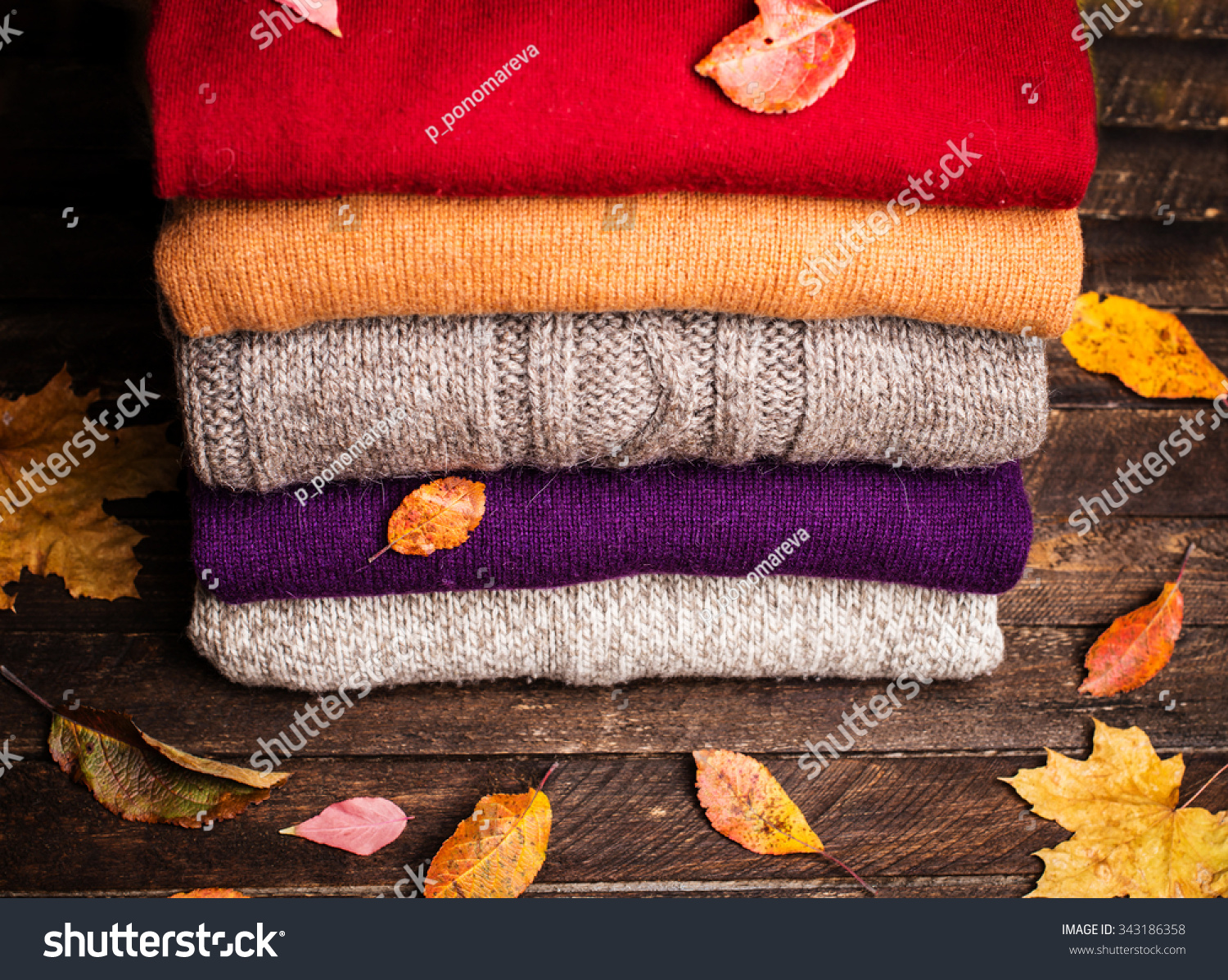 Pile of knitted winter clothes on wooden background covered with autumn leaves  knitwear  space for text. Stack of knitted sweaters and cardigans.
