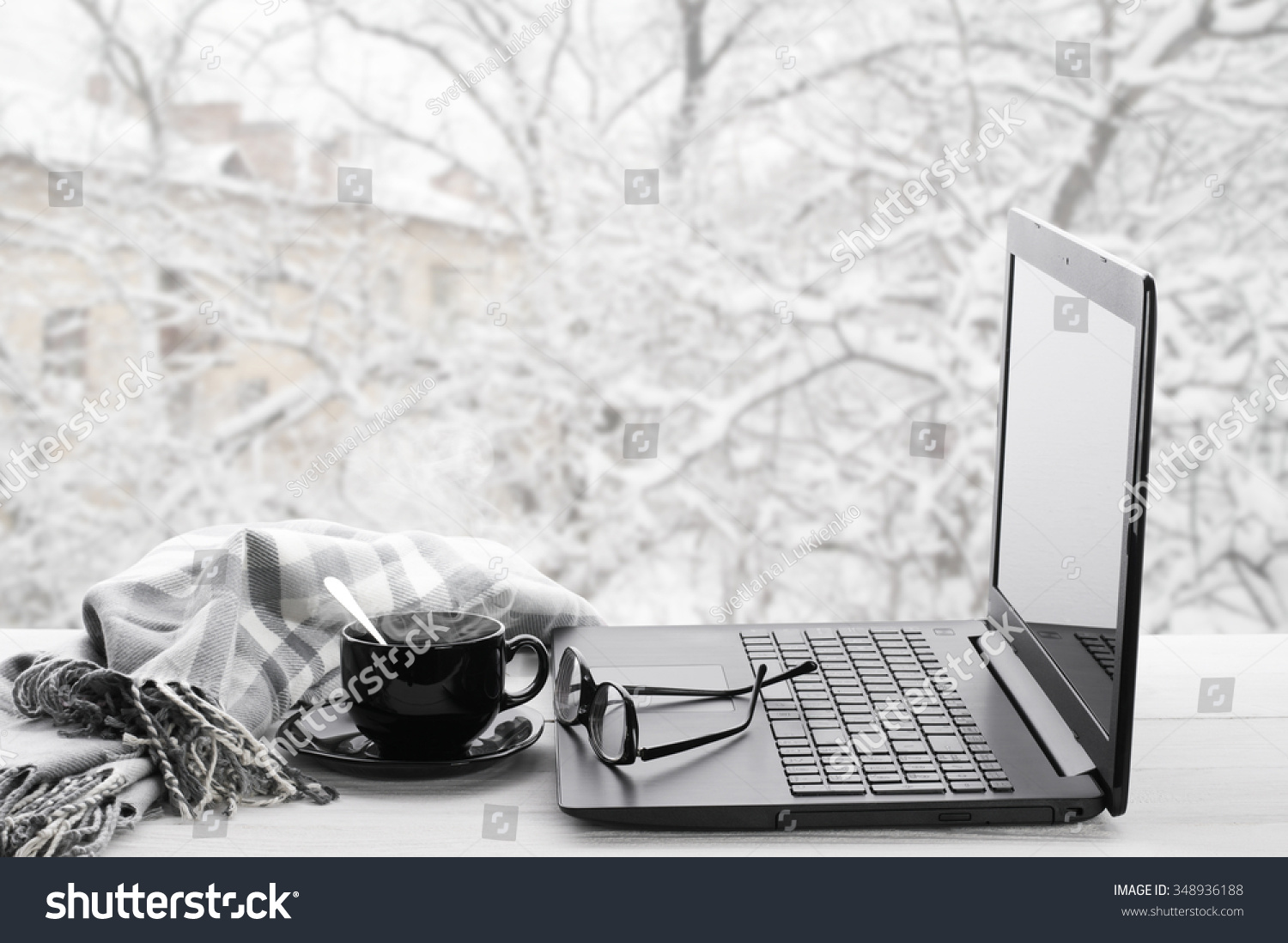 Cozy winter still life: laptop glasses cup of hot coffee and warm plaid on windowsill against snow landscape from outside.