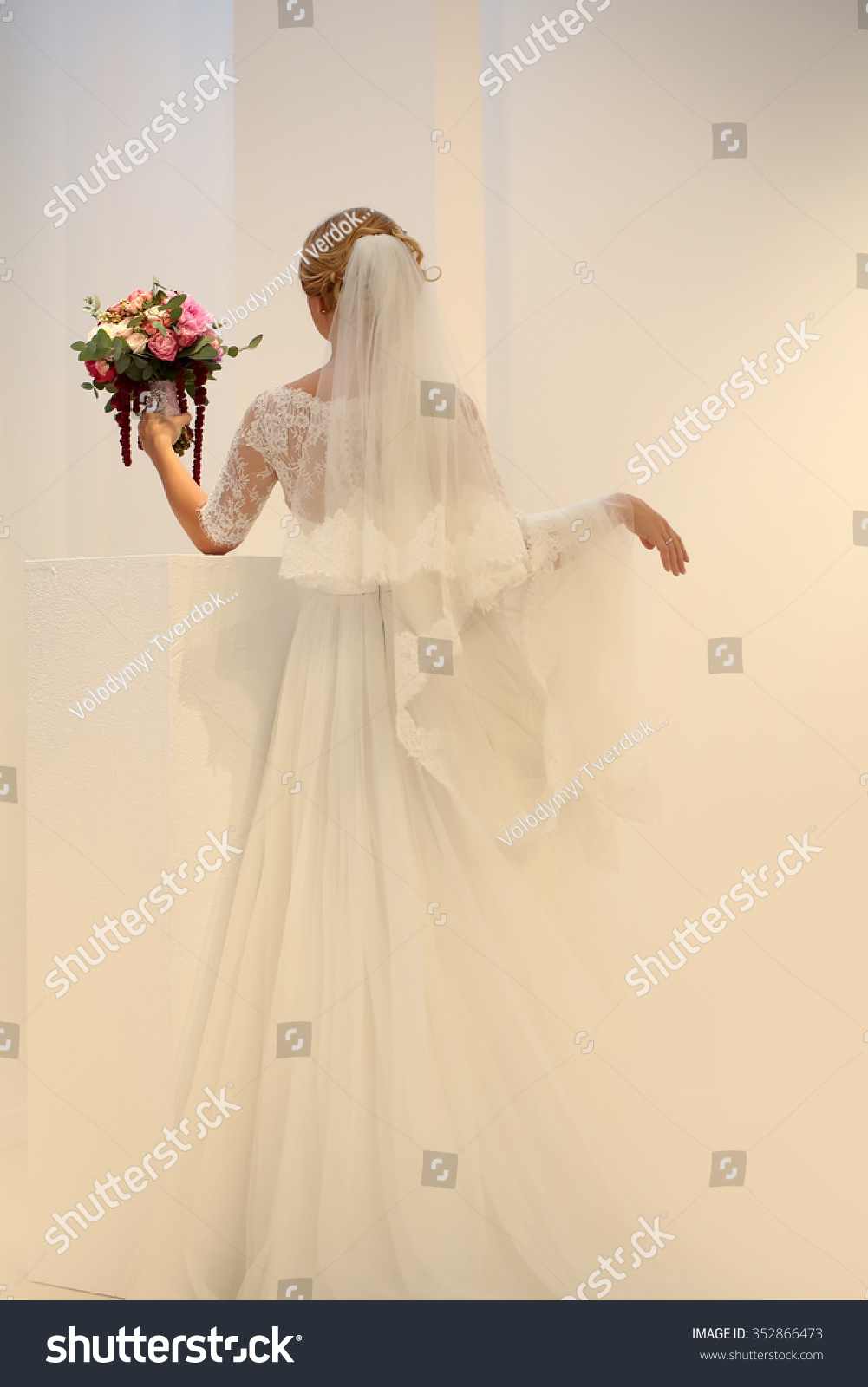 Photo of young bride full length back view wearing long lace white wedding dress and bridal veil posing with beautiful decorated bouquet for ceremony in hand on pastel background  vertical picture
