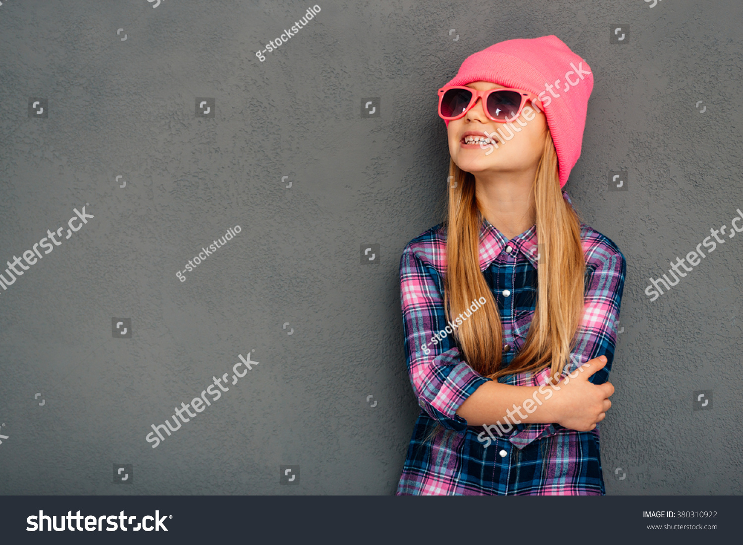Stylish and cute. Cheerful little girl in sunglasses keeping arms crossed and looking up with smile while standing against grey background