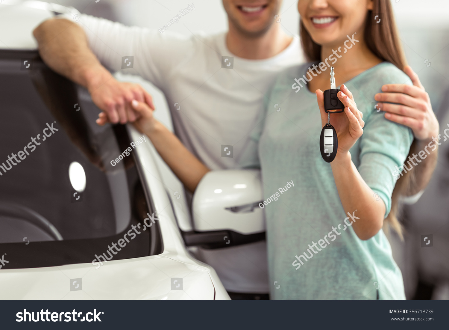 Beautiful young couple is smiling and looking at camera while leaning on their new car in a motor show. Woman is holding car keys close-up