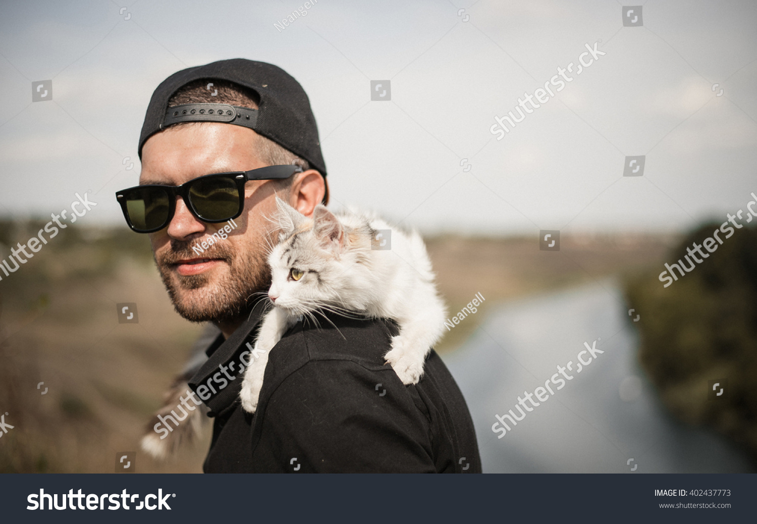 a bearded man in a black baseball cap shirt and shorts playing with white fluffy cat on top of a cliff on the background of the river and the forest