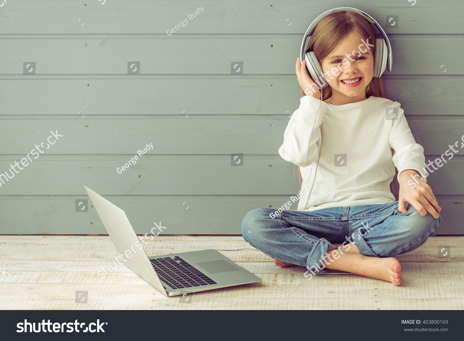 Pretty little girl in headphones is listening to music using a laptop  looking at camera and smiling  against gray background