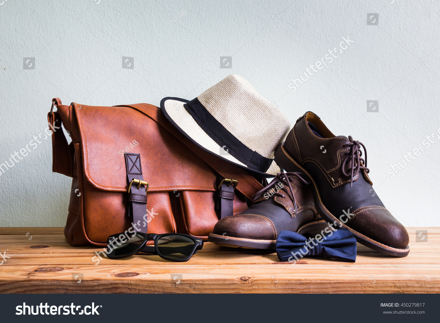 Men's accessories with brown leather bag  brown shoes  classic hat  sunglasses and blue bow tie on wooden table over wall background