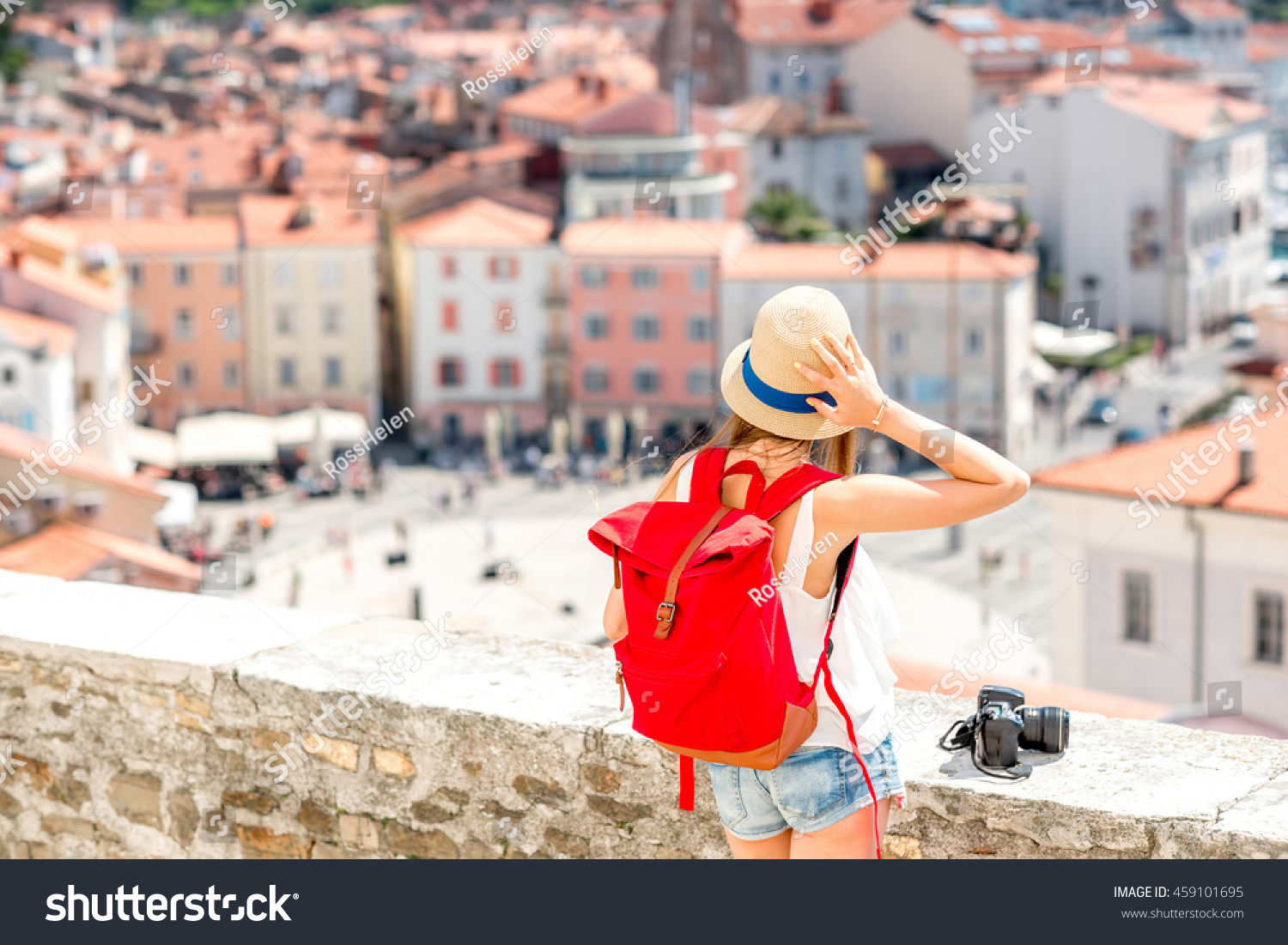 Young female traveler with hat red backpack and photo camera enjoying the view on Piran old town. Back view with copy space. Traveling in Slovenia